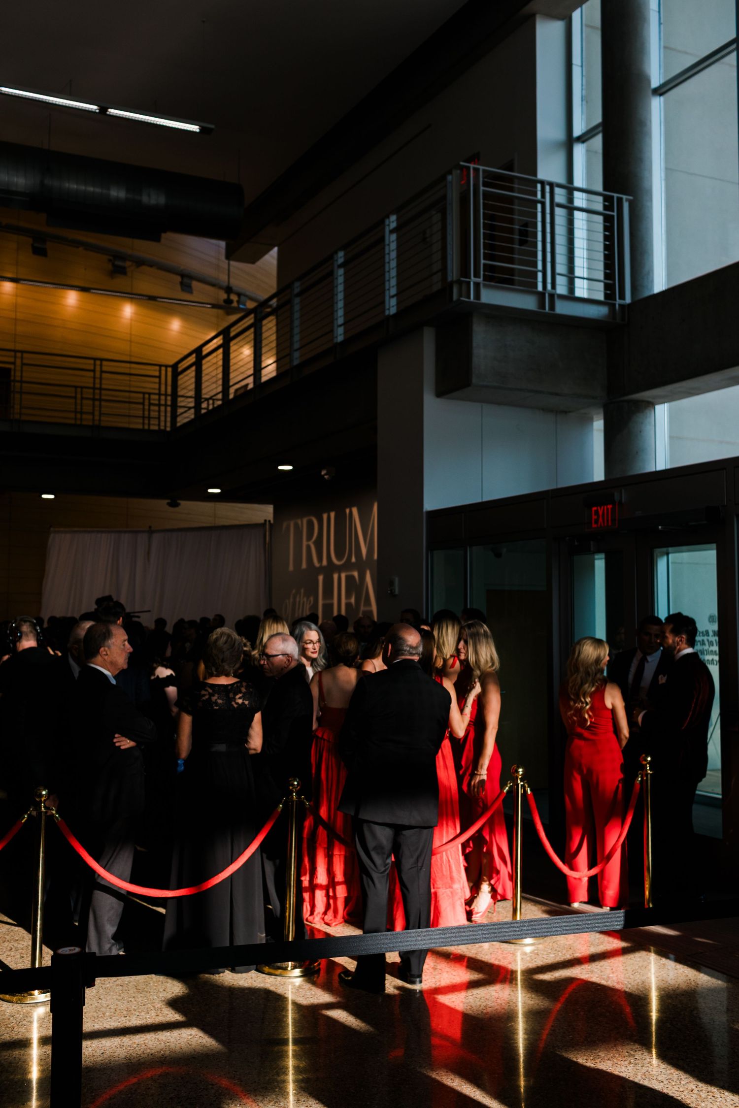 People in formal black and red attire gather in a modern indoor space with dramatic lighting and rope barriers.