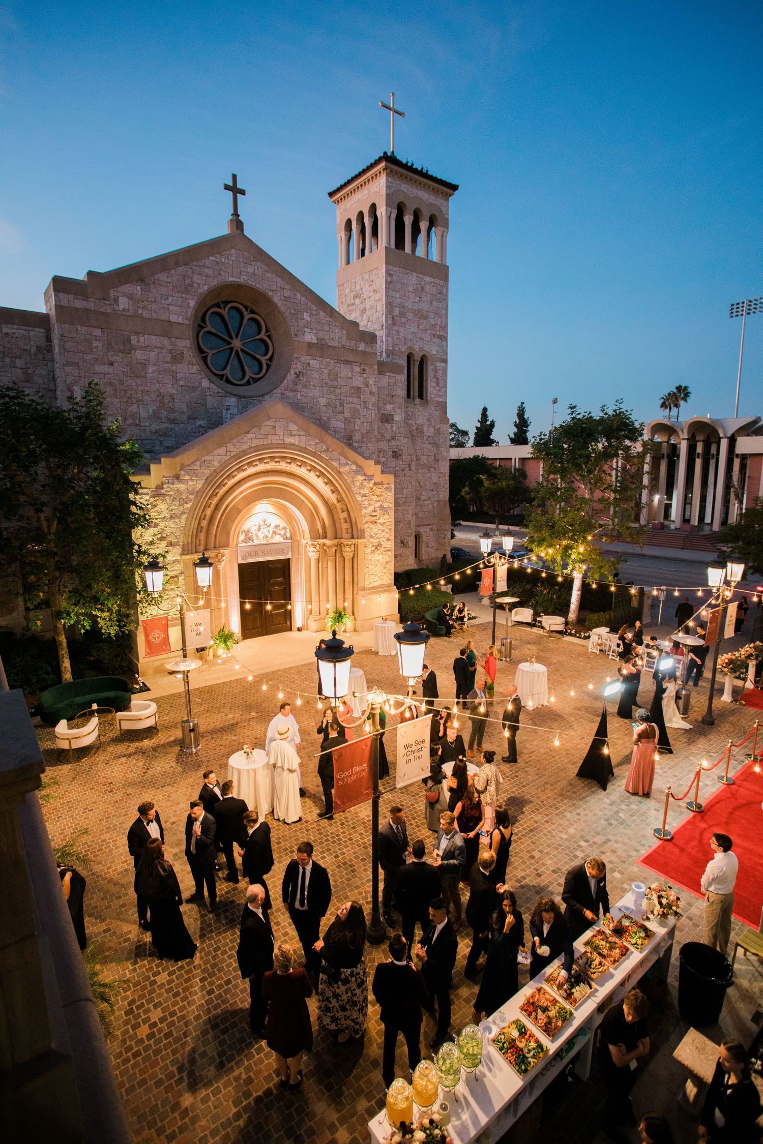 Evening wedding ceremony outside illuminated church with guests gathering on stone courtyard at sunset.