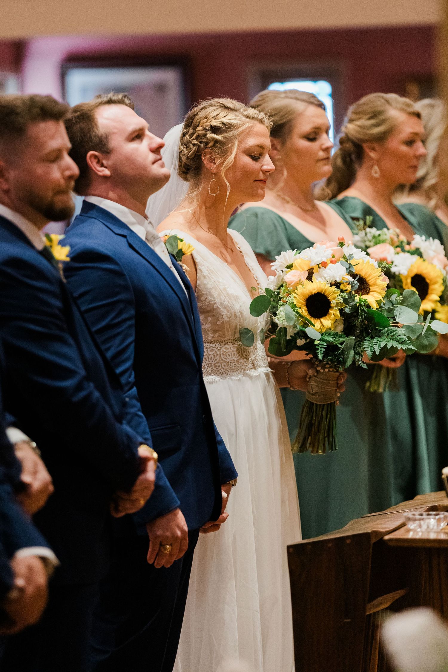Bride and groom stand at the altar holding hands, flanked by groomsmen and bridesmaids in green dresses with sunflower bouquets.