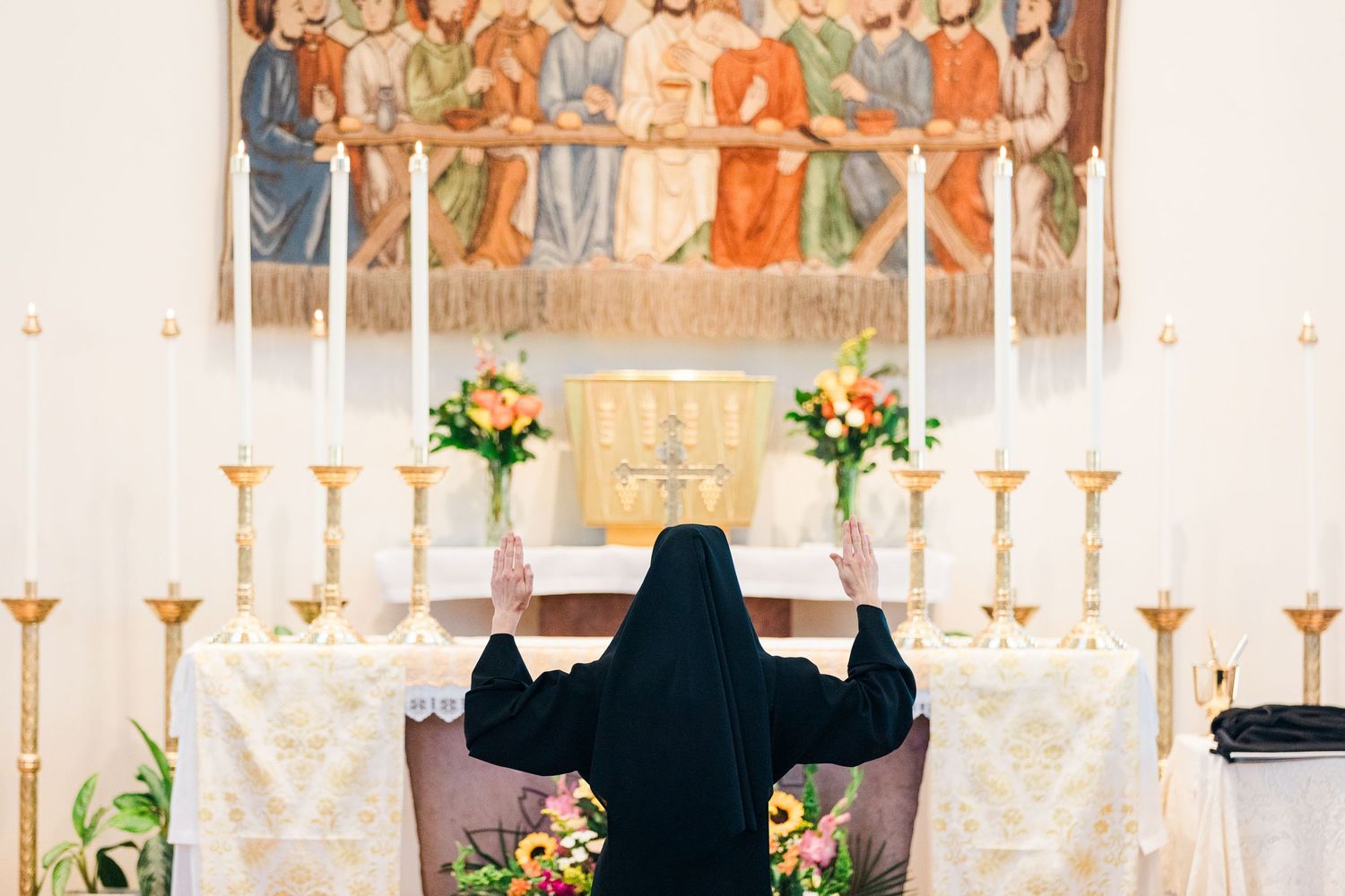 A nun prays at an ornate church altar adorned with candles, flowers, and a religious tapestry depicting saints.