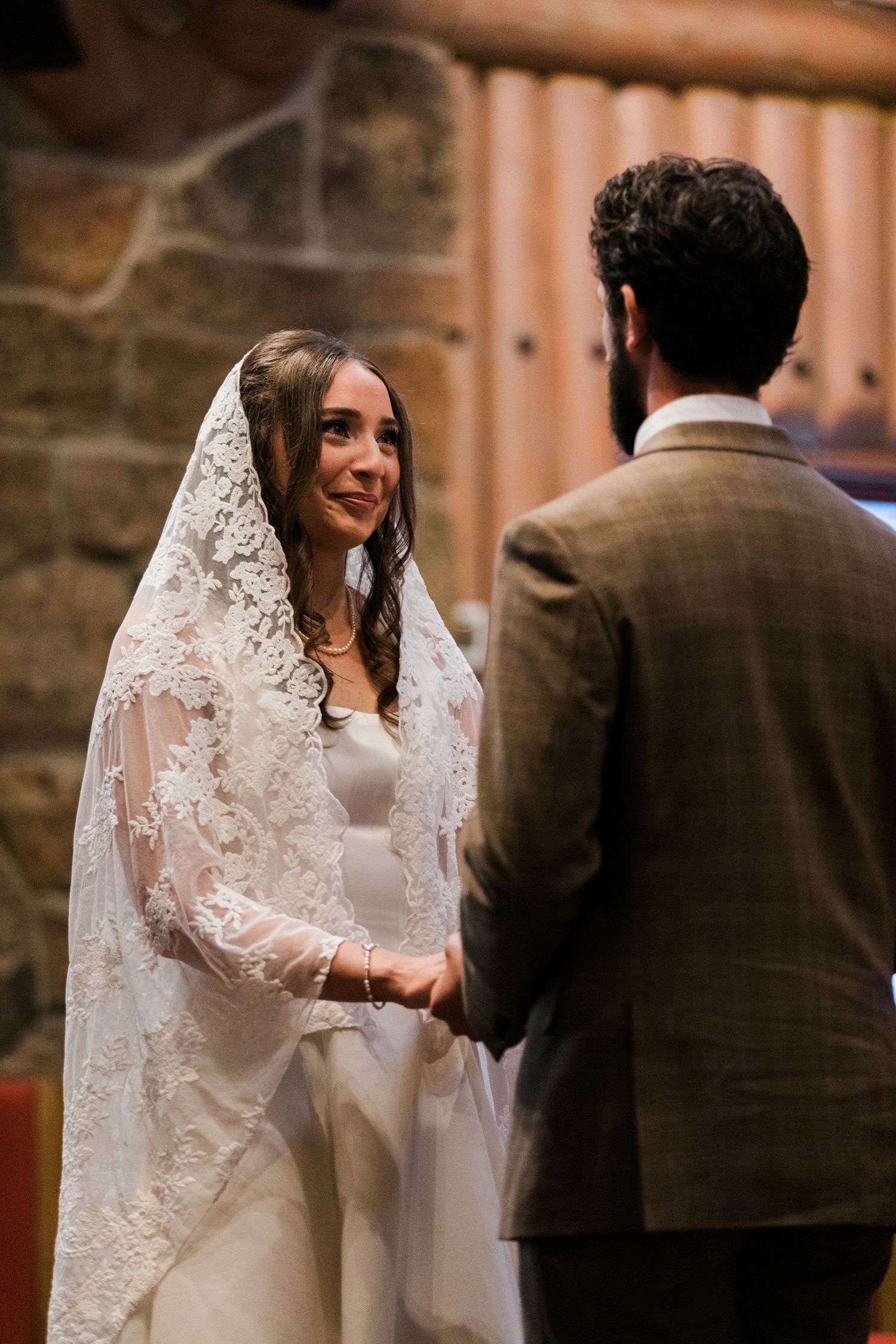 Bride in lace veil smiles at groom during wedding ceremony in rustic stone chapel.