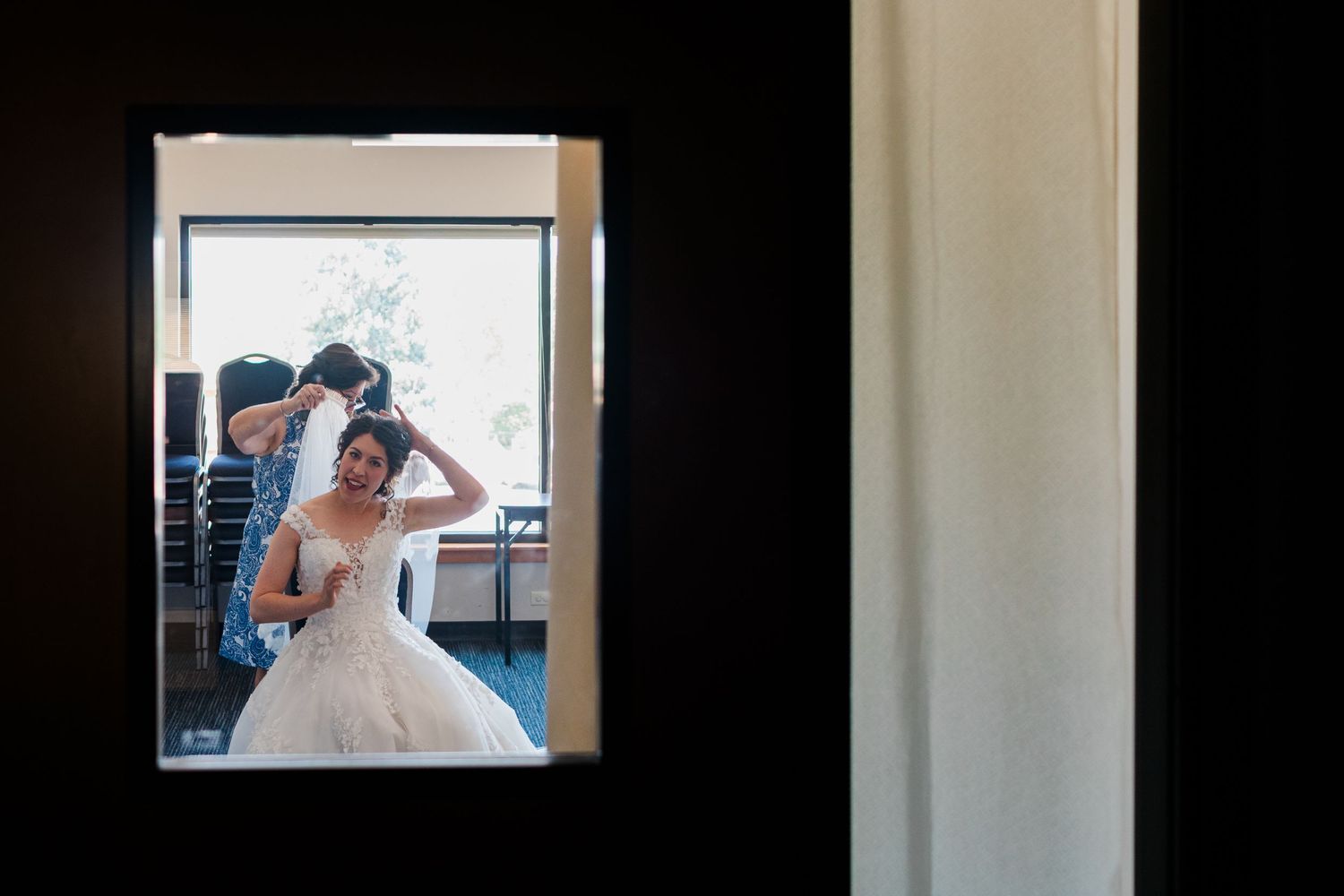 Bride in white wedding gown getting hair styled by assistant, reflected in mirror during bridal preparation.