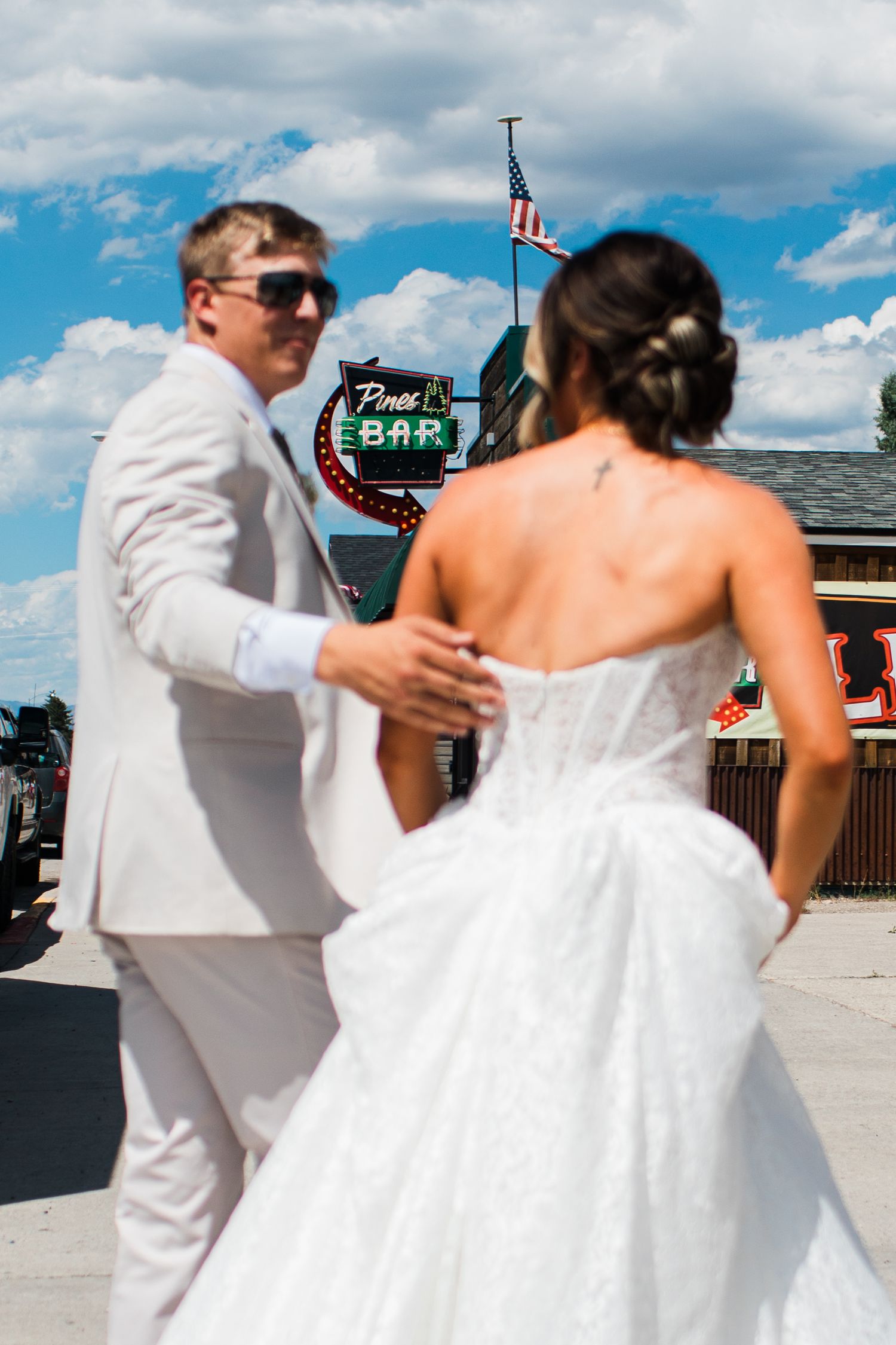 Bride and groom in white attire share a moment outside a bar with an American flag on a sunny day.