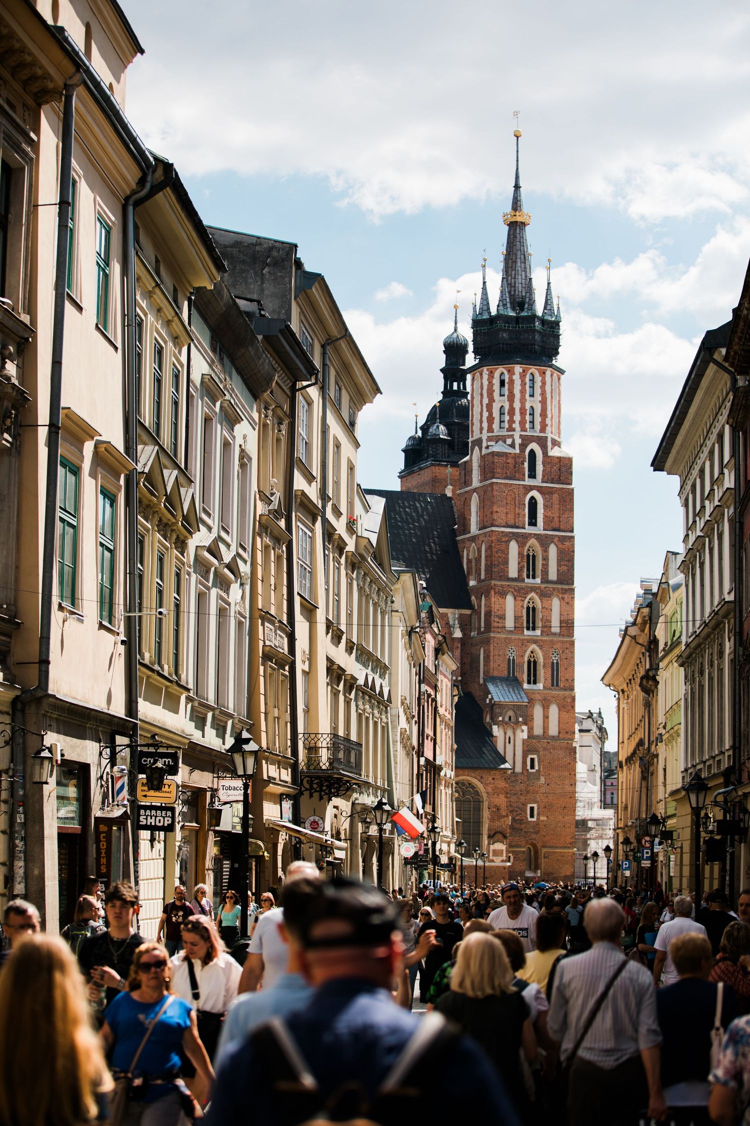 Crowded Florianska Street in Krakow with St. Mary's Basilica Gothic towers rising above historic buildings on a sunny day.