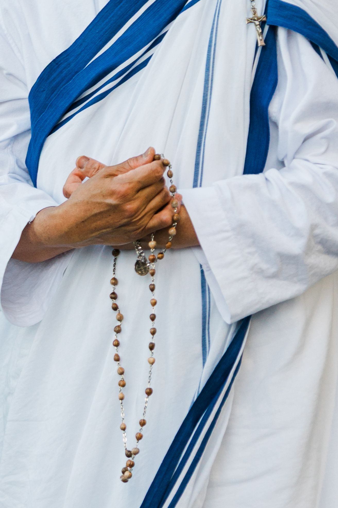 Catholic nun in white and blue habit holds wooden rosary beads while praying, close-up of clasped hands.