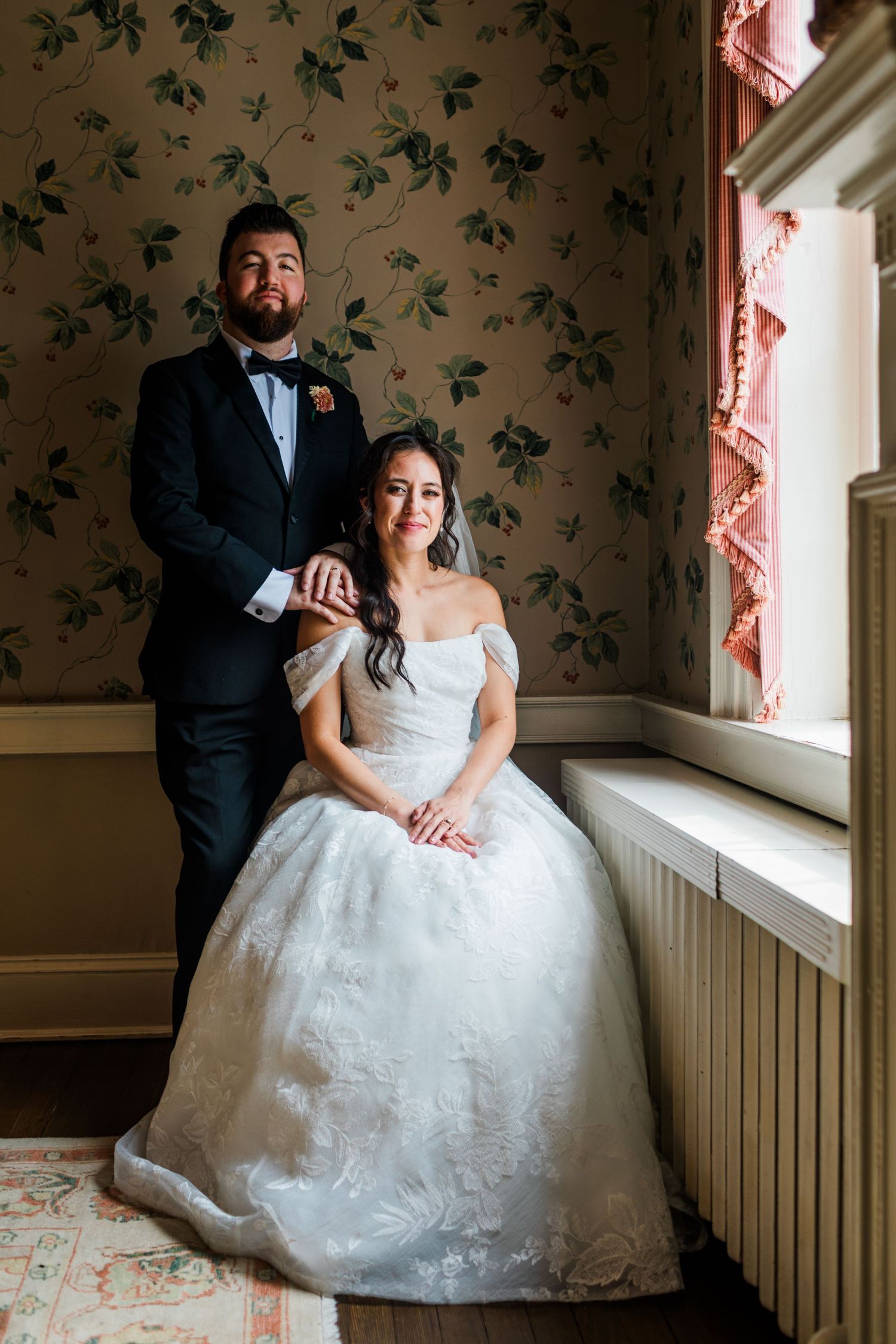Bride in white off-shoulder ball gown sits by window as groom in black tuxedo stands beside her in floral wallpapered room.