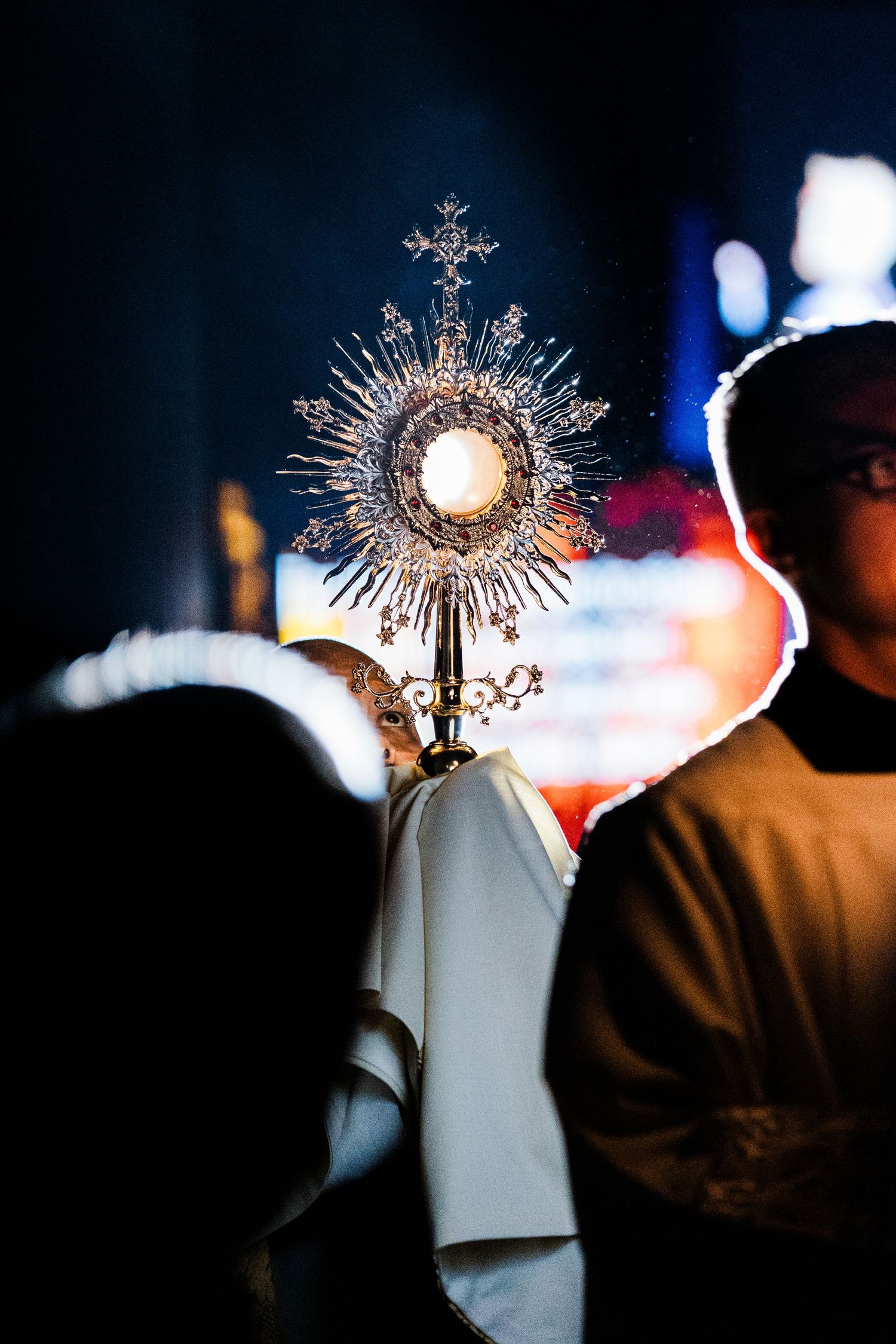 A golden monstrance with ornate sunburst design glows against a dark background during Catholic worship.