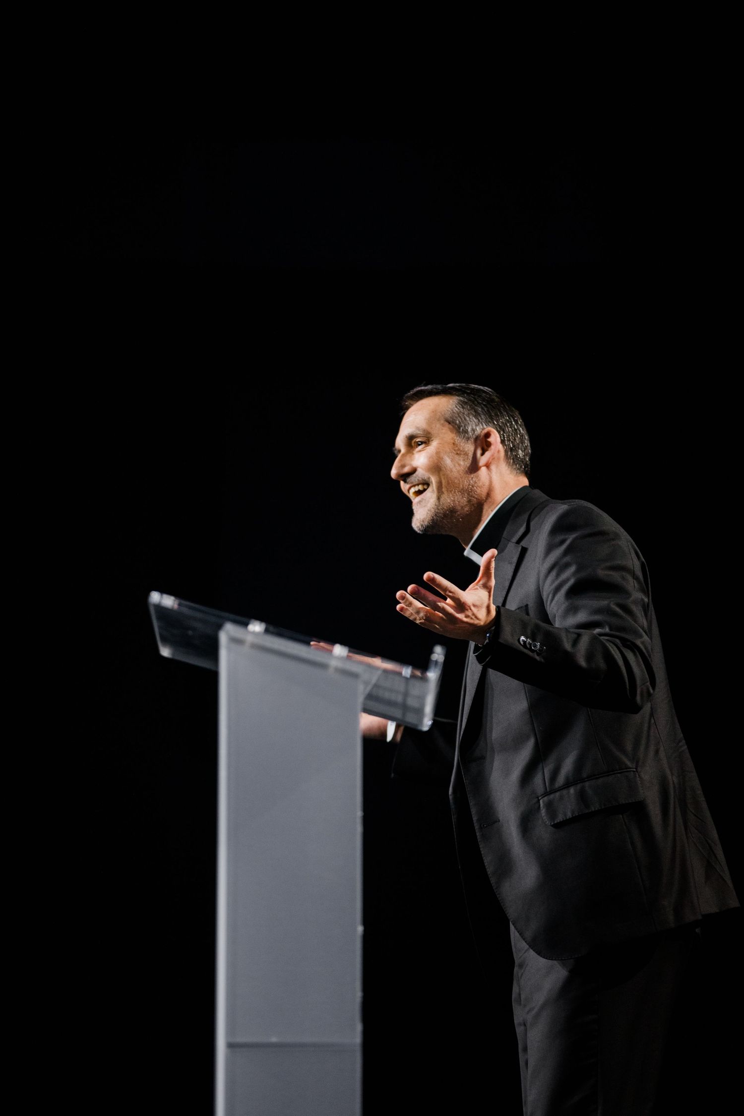 A speaker in a black suit gestures expressively while presenting at a podium against a dark background.