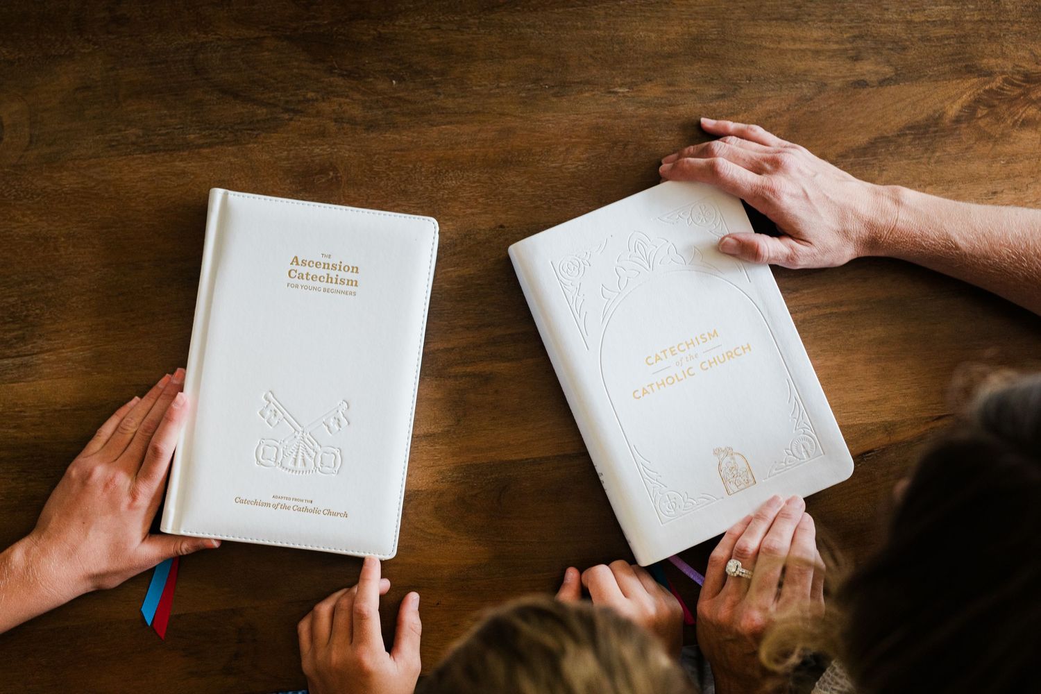Two people holding white hardcover books with ribbon bookmarks over a wooden table.