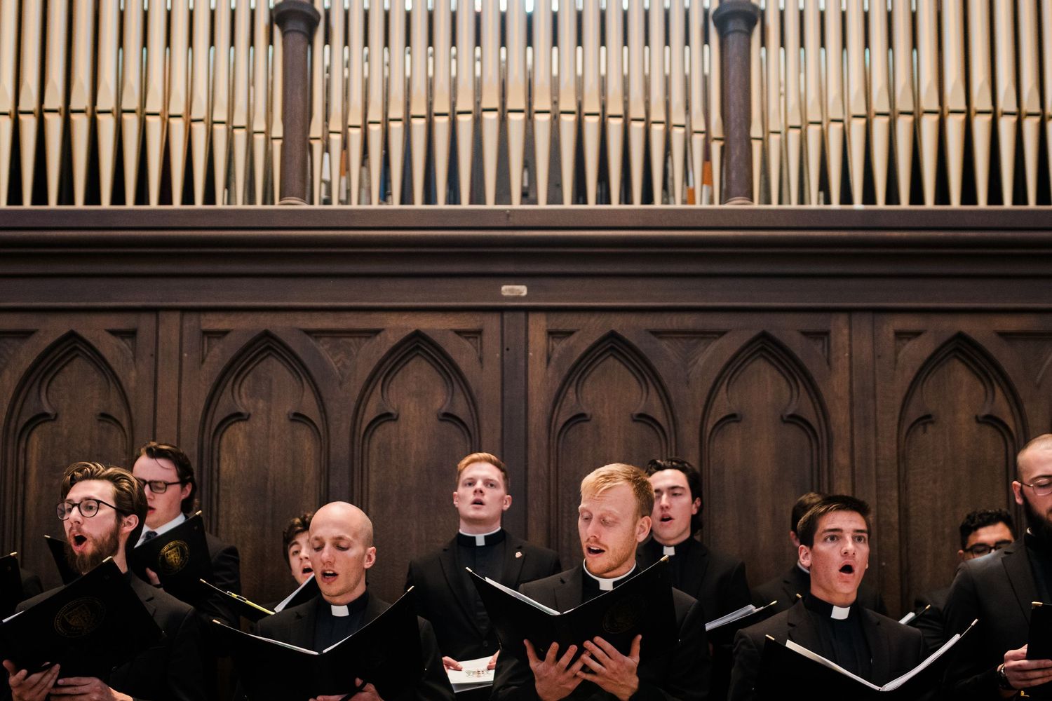 A choir performs in formal black attire while standing in rows inside a church or concert hall.