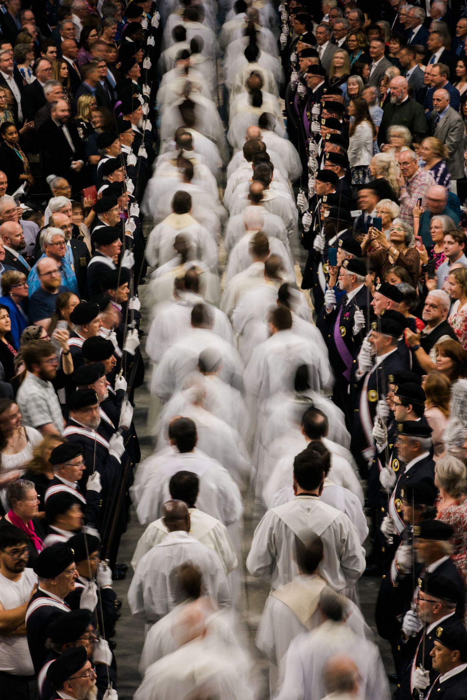A large crowd at a formal event with people in dark suits and others wearing white robes and veils gathered together.