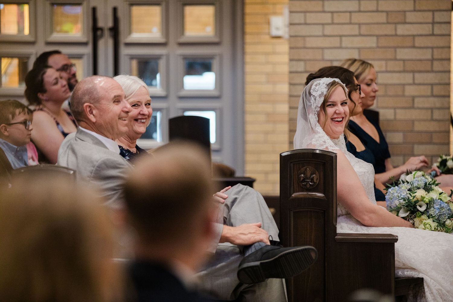 Bride in white veil smiles during wedding ceremony while seated in church pew with guests.