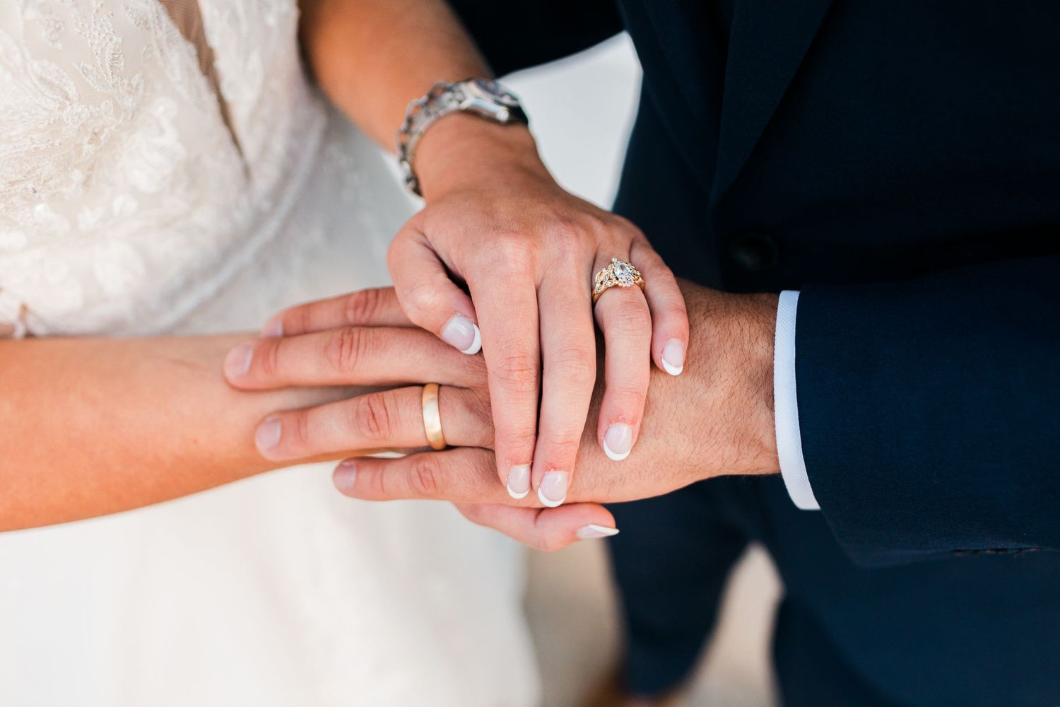 Close-up of newlyweds hands showing wedding rings, bride in white dress and groom in navy suit.