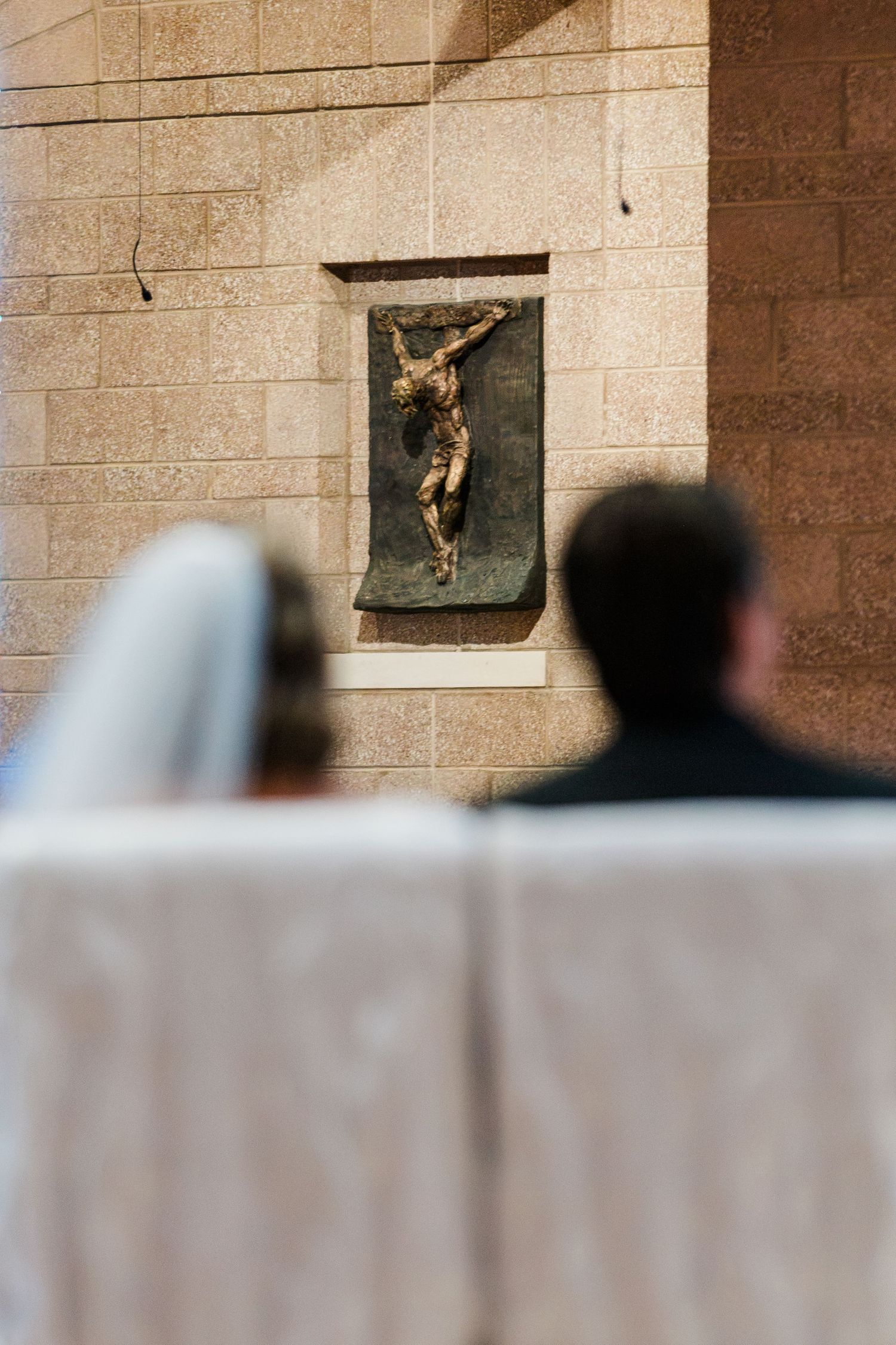 Bride and groom kneeling before a bronze crucifix sculpture on stone wall during a Catholic wedding ceremony.