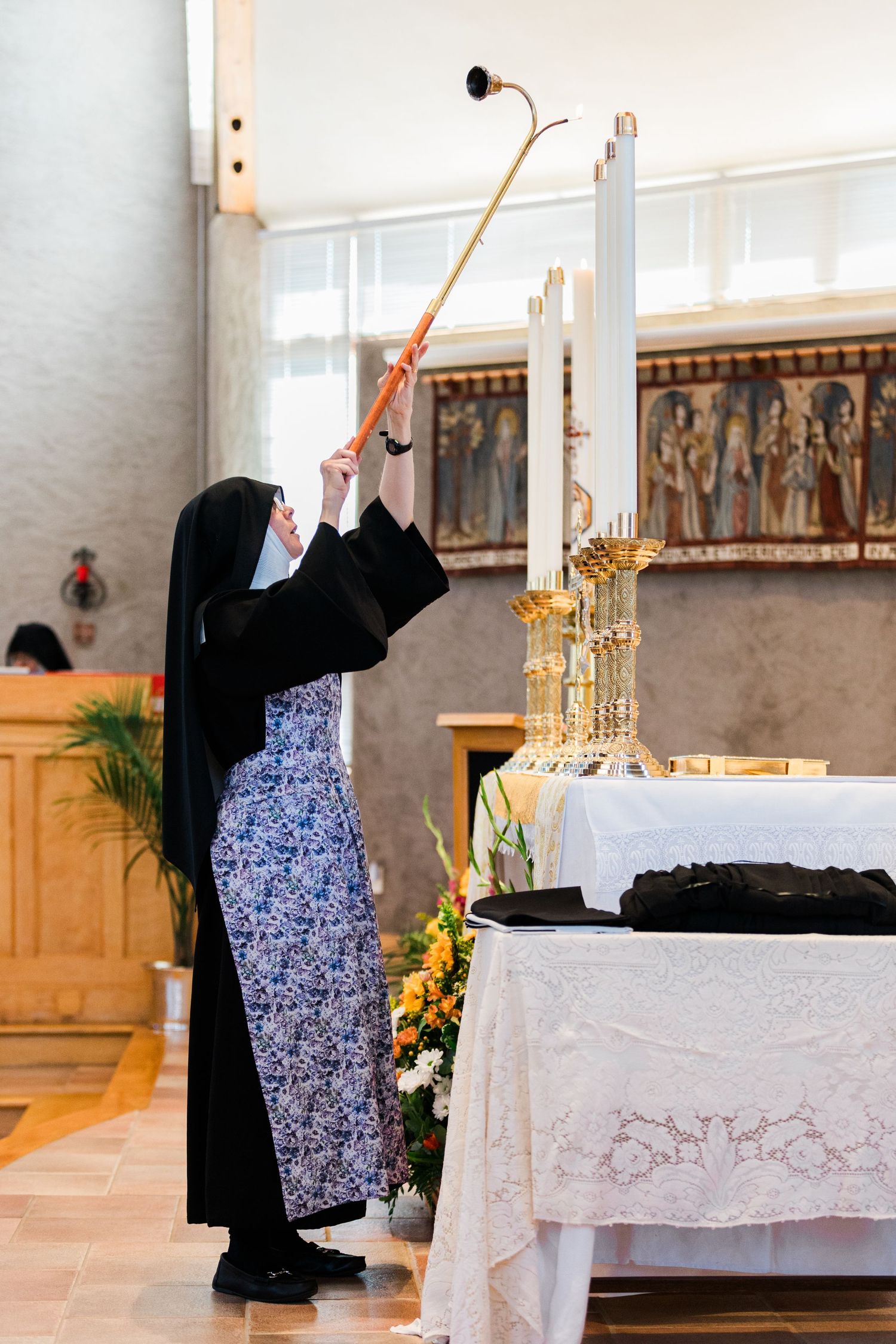 A nun in traditional habit uses a long candle snuffer at a church altar adorned with flowers and candles.