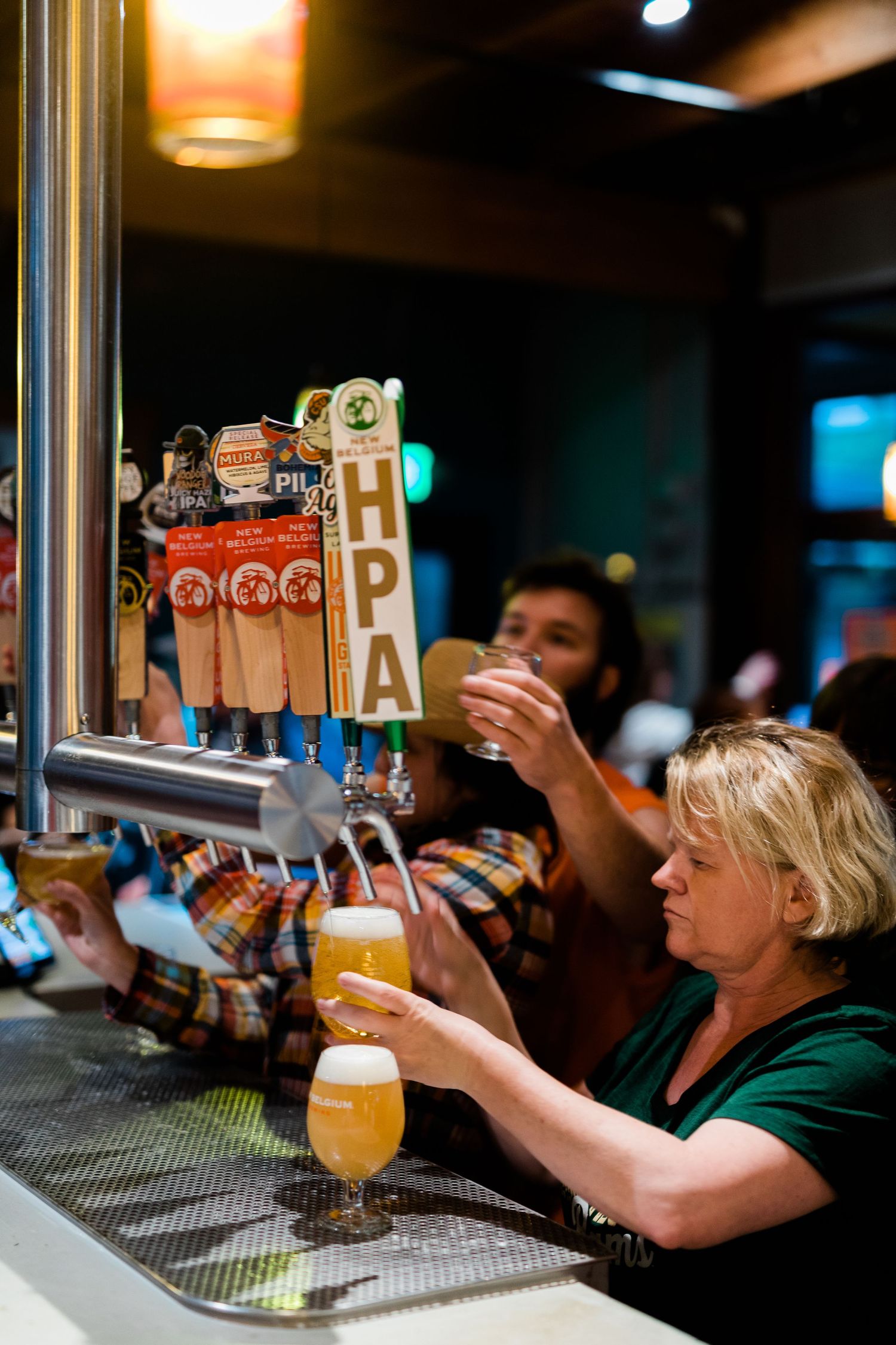 A row of craft beer taps at a bar with patrons enjoying drinks in the background.