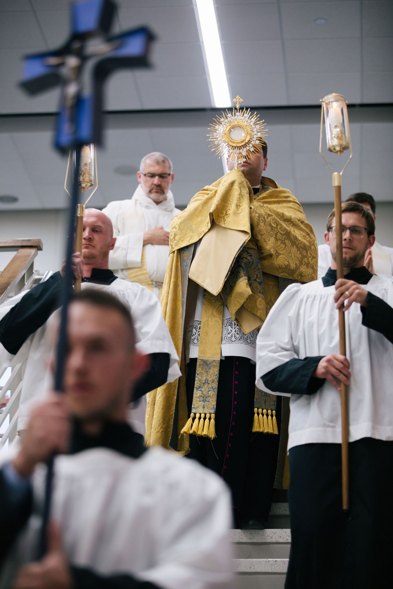 Catholic priest in gold vestments holds monstrance during Eucharistic adoration ceremony with altar servers in white robes.