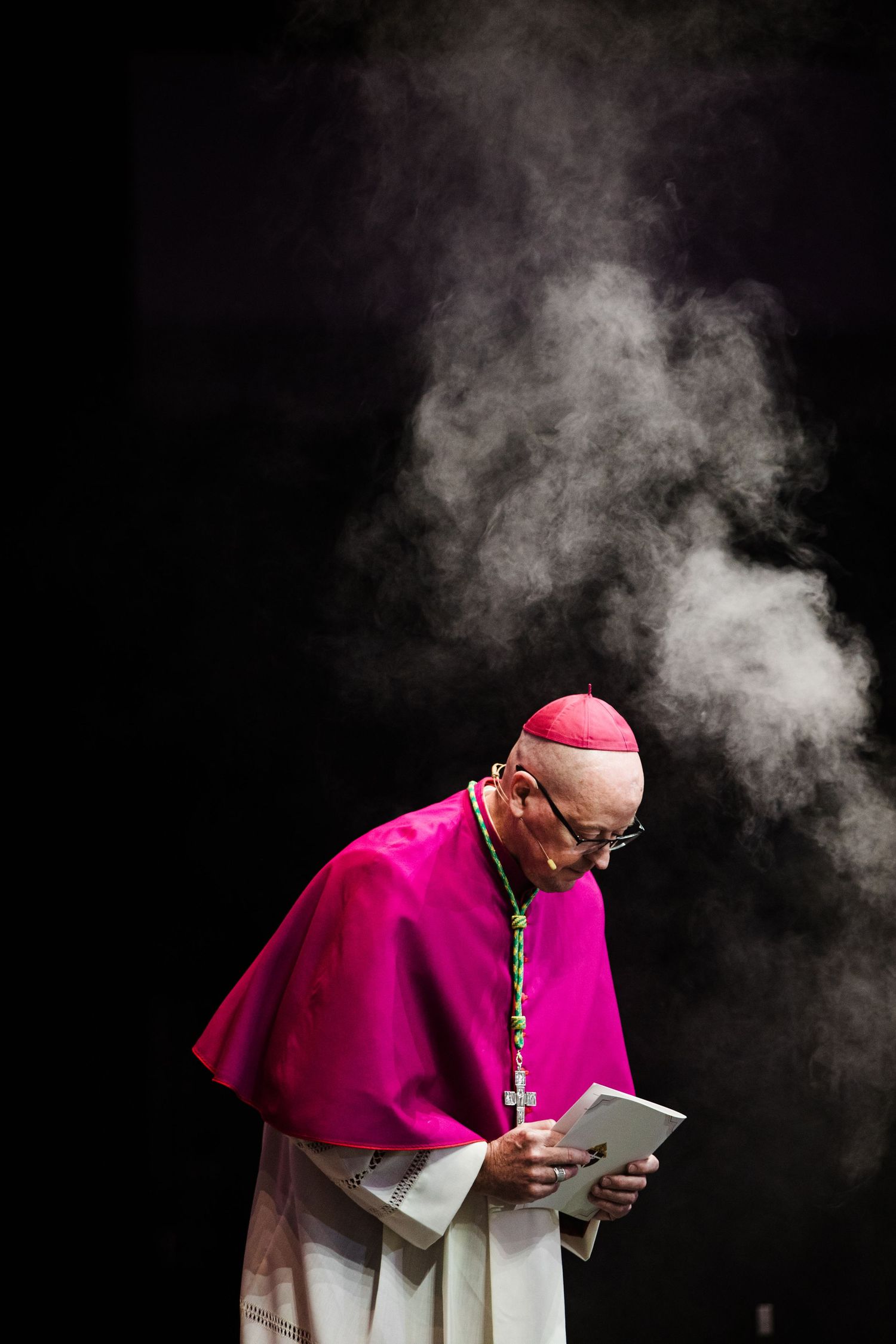 Catholic bishop in magenta vestments reads from a tablet during a ceremonial mass with incense smoke rising behind him.