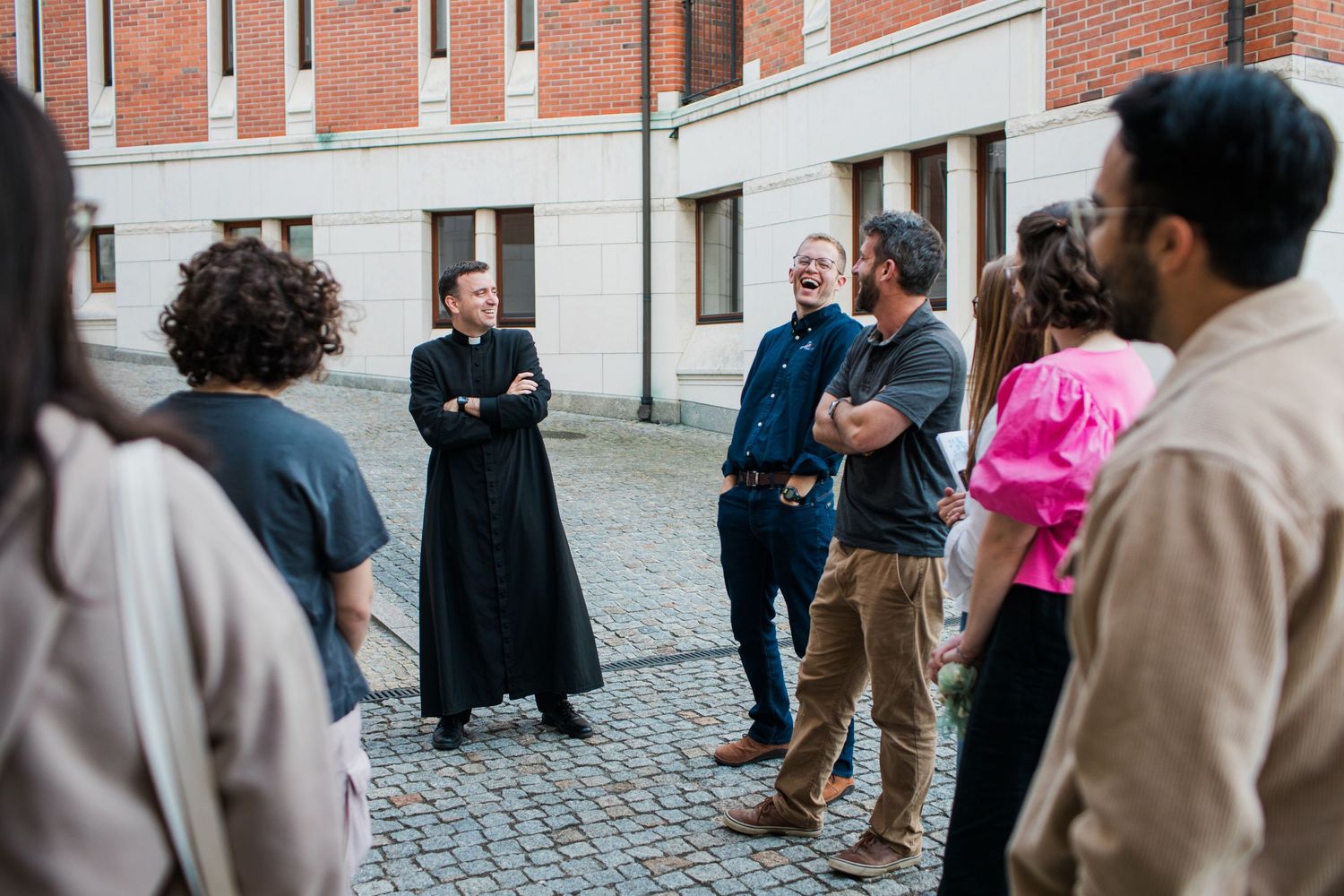 A Catholic priest in black cassock laughs with a diverse group of visitors during an outdoor tour of a brick building.