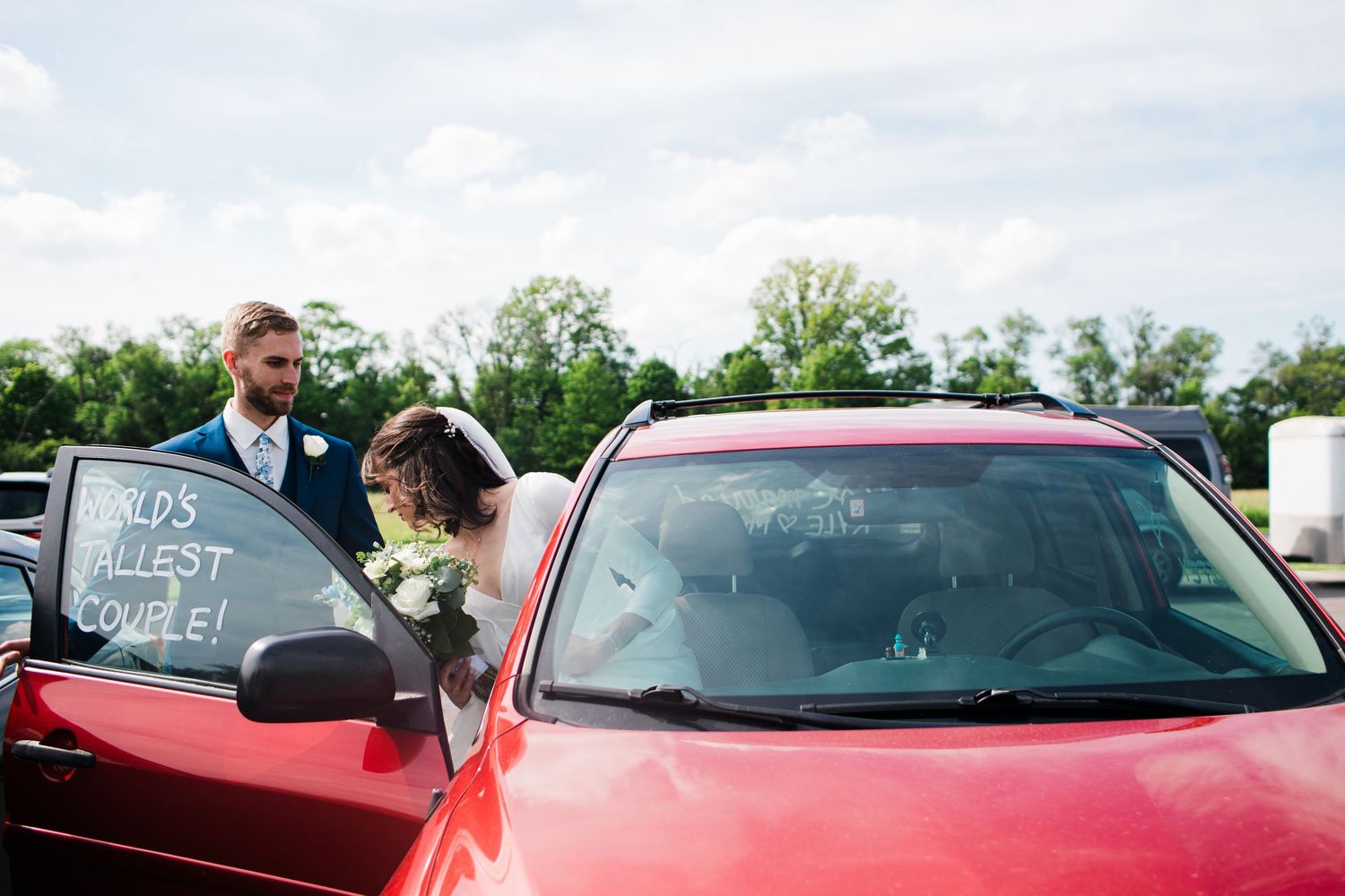Newlywed couple in wedding attire standing beside a red car with World's Luckiest Couple written on the window.