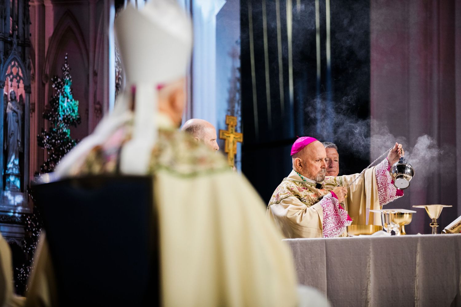 A Catholic bishop in pink robes performs a religious ceremony at an altar with incense smoke rising in a cathedral.