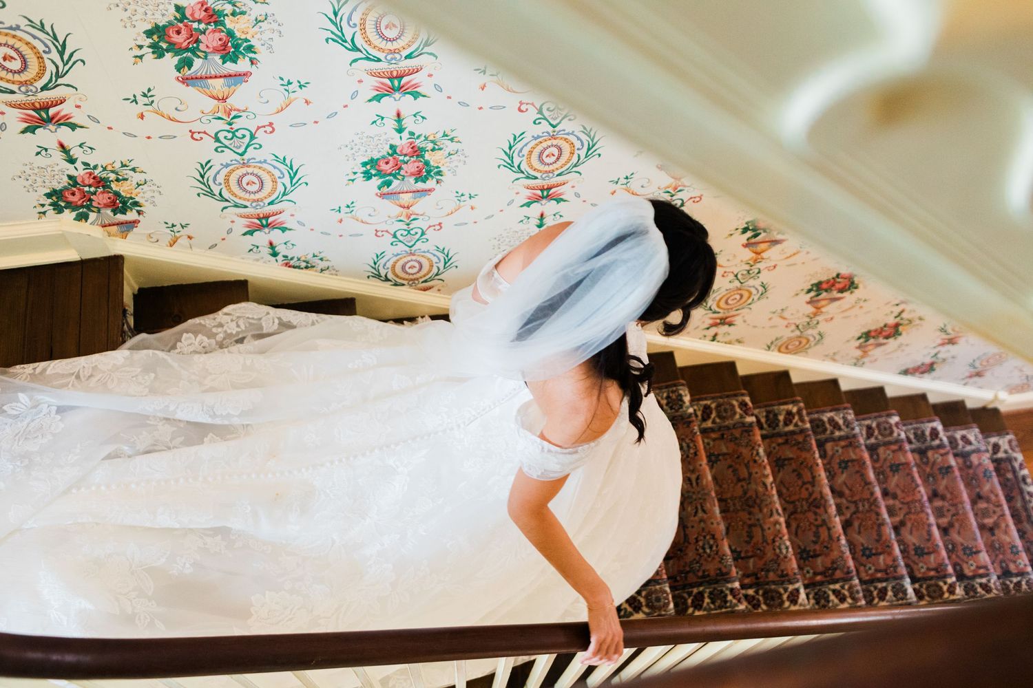 Bride in white wedding gown and veil descending elegant staircase with floral wallpaper.