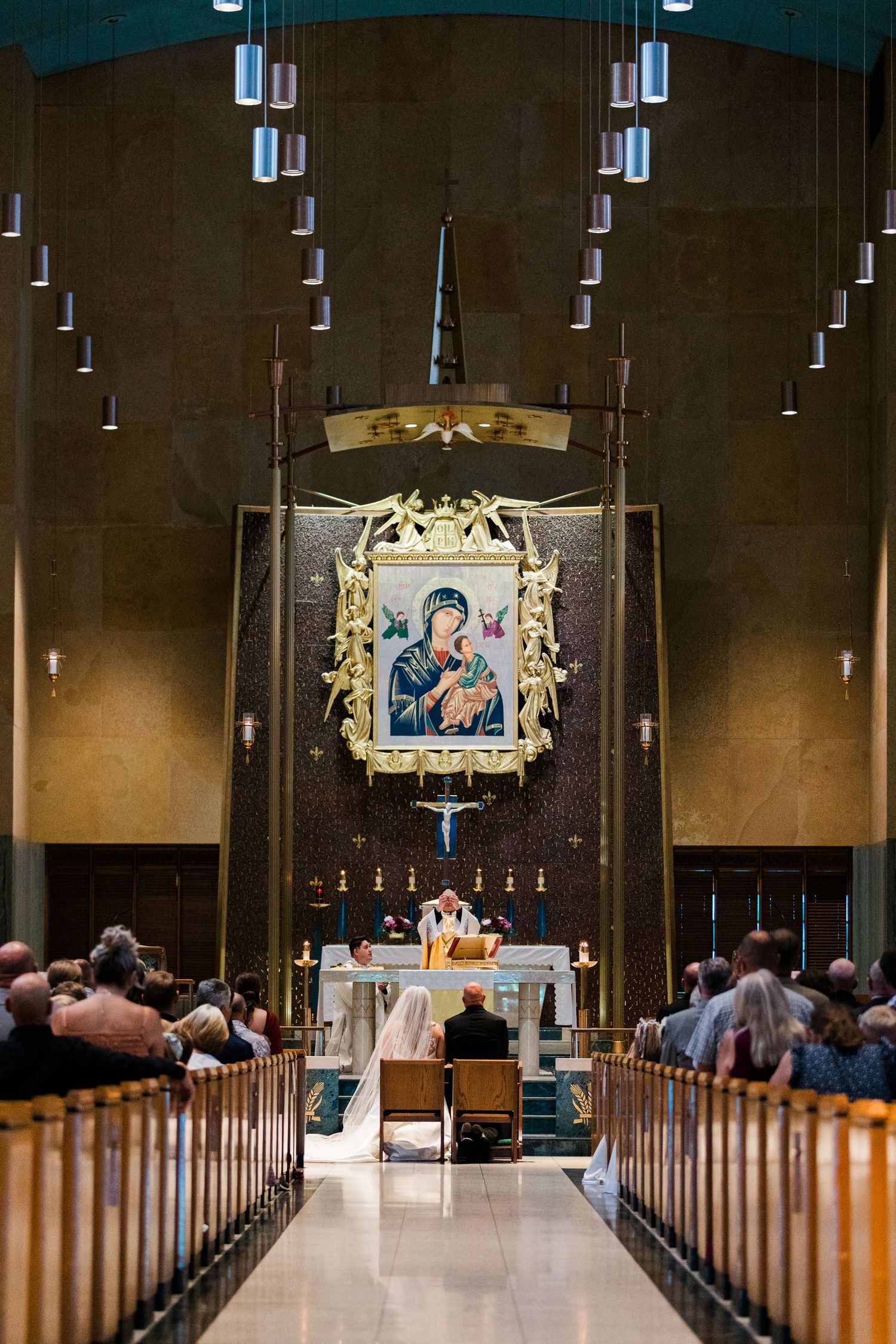 Catholic church wedding ceremony with couple at ornate altar beneath Virgin Mary icon, surrounded by wooden pews of seated guests.