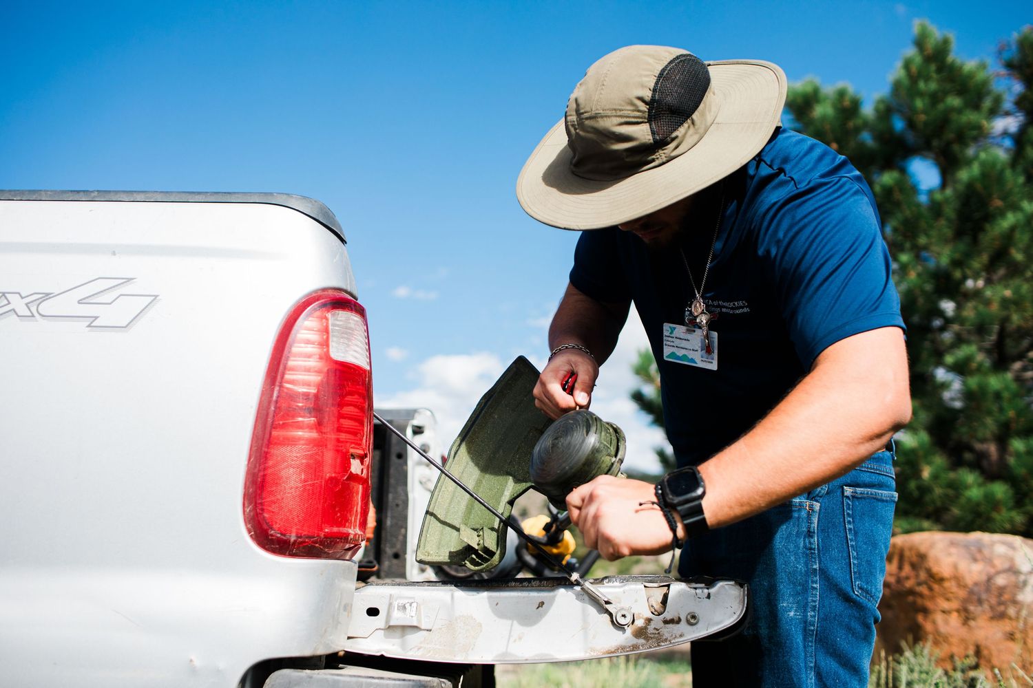 Man in hat and blue shirt unpacking equipment from a truck bed outdoors on a sunny day.