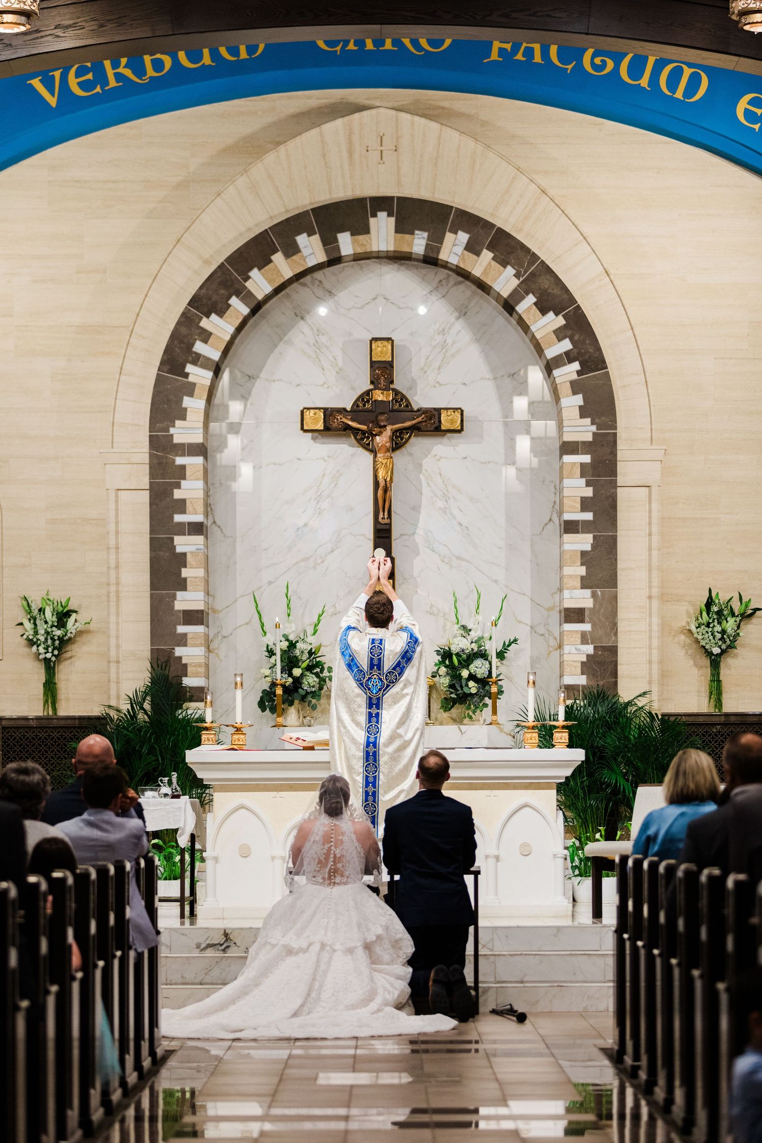 Bride and groom kneel at altar during Catholic wedding ceremony with priest and crucifix.