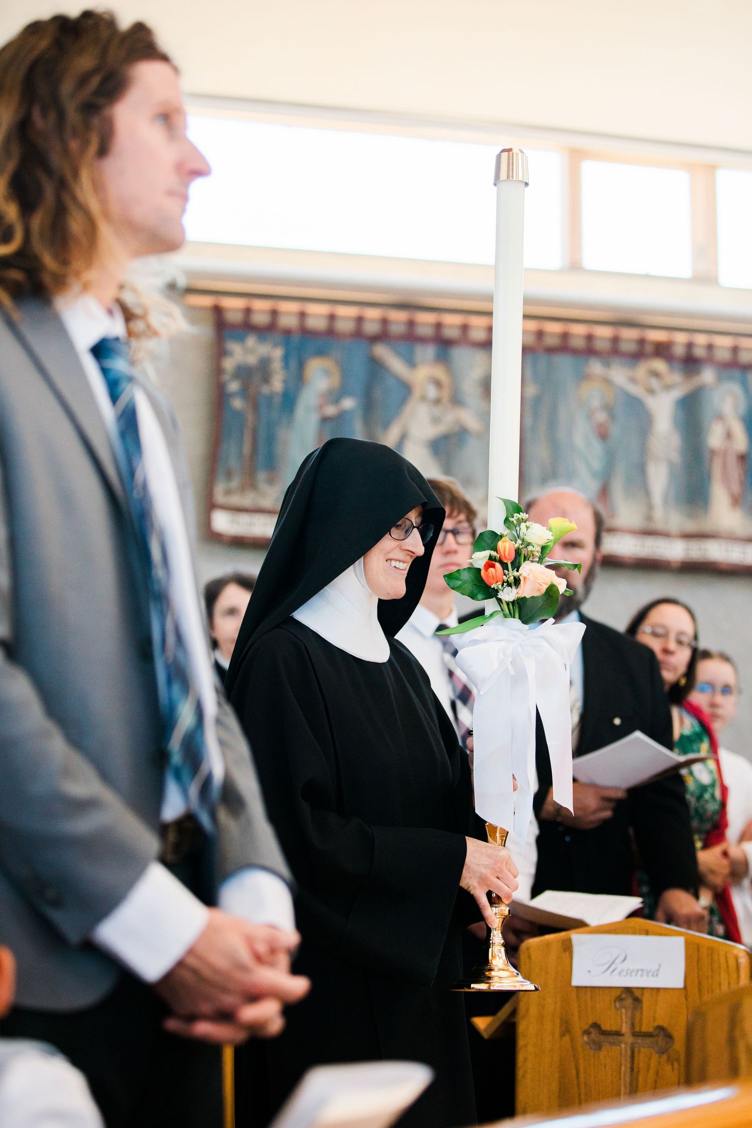 A smiling nun in black habit holds a candle during a church ceremony with attendees in formal attire.