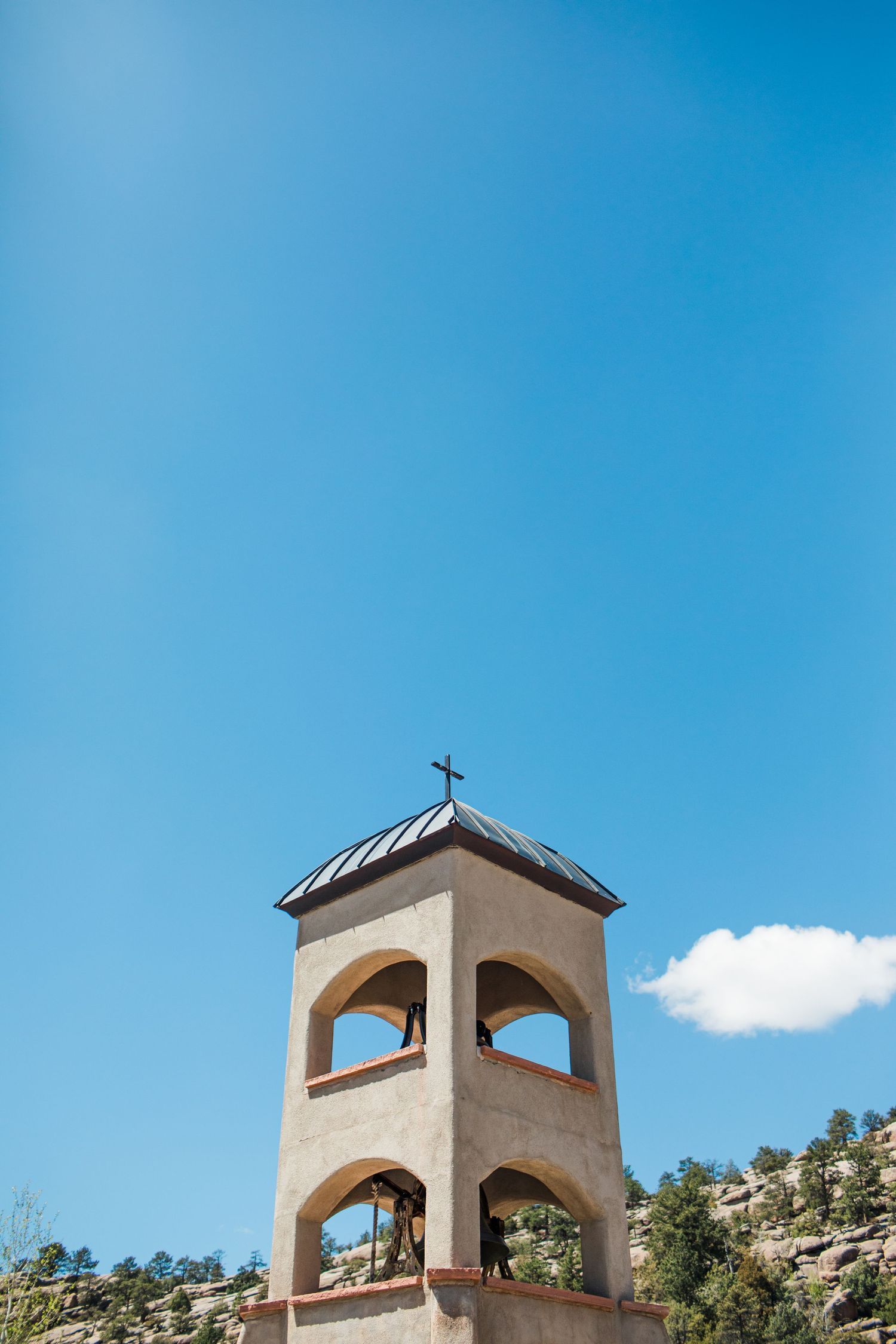 Adobe bell tower with cross atop arched openings against a bright blue sky, surrounded by rocky hills and pine trees.