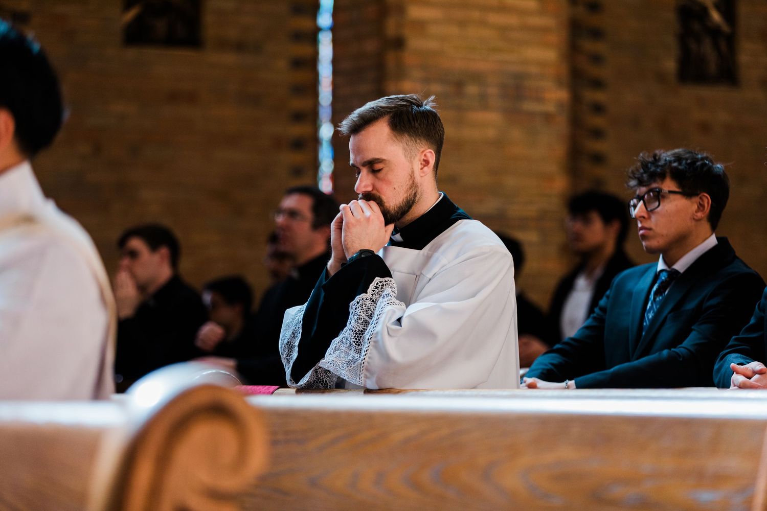 Young Catholic deacon prays reverently during a church ceremony, hands clasped near his face in a pew.