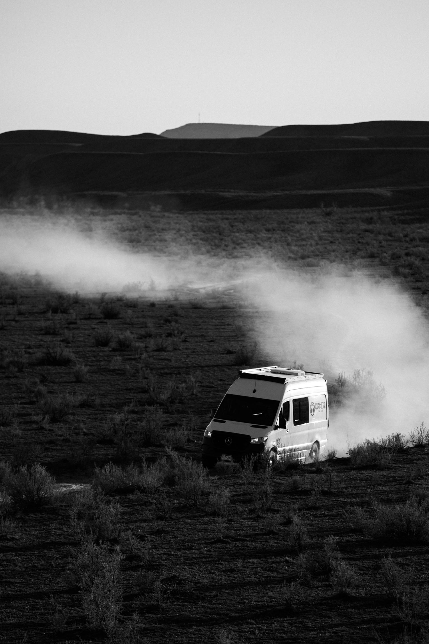 White SUV kicks up dust while driving through a desert landscape in dramatic black and white photography.