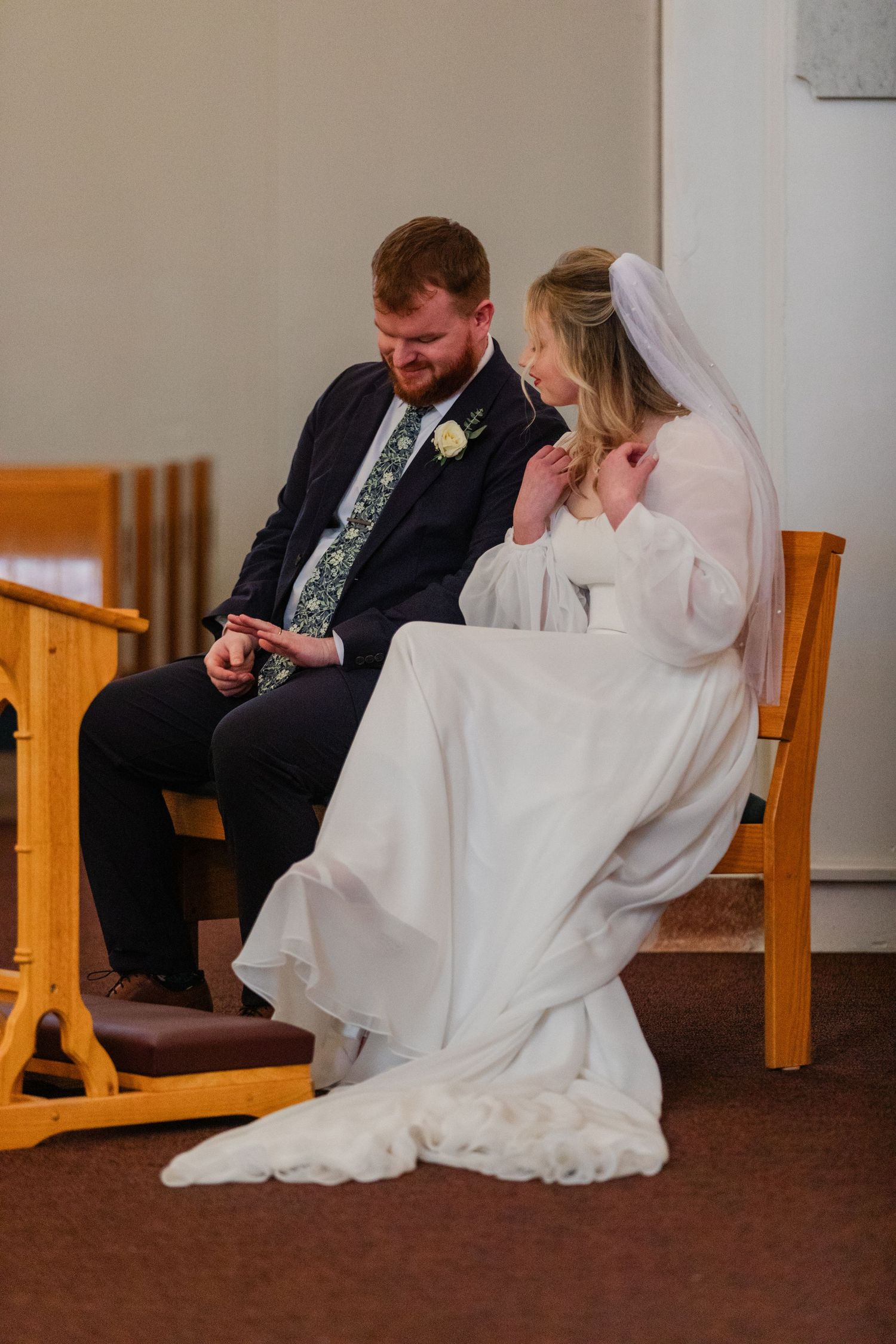 A couple sits together on wooden chairs in a quiet moment during their wedding ceremony.