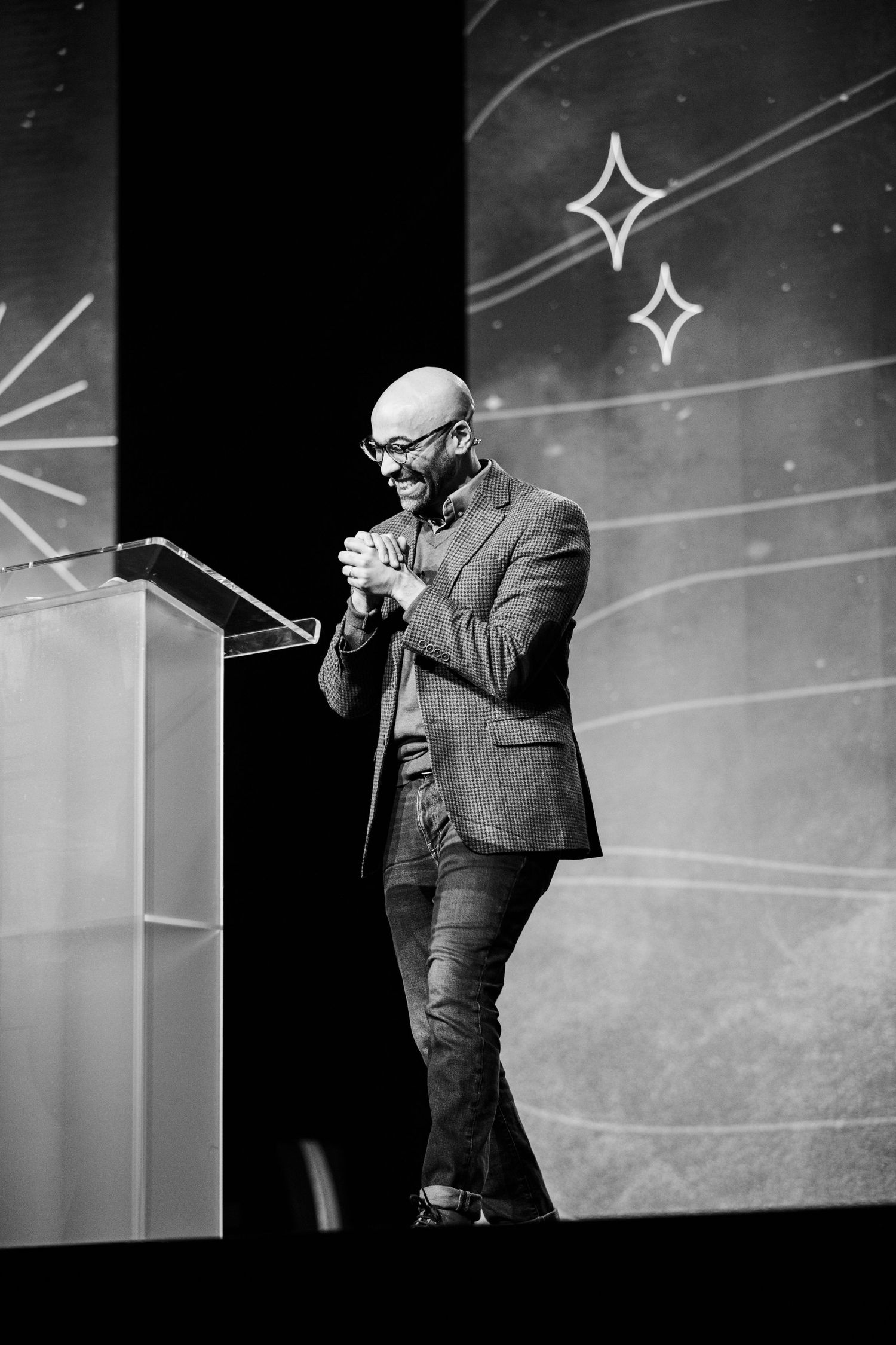 A speaker in a grey blazer and jeans stands at a podium on stage with decorative stars on the backdrop.
