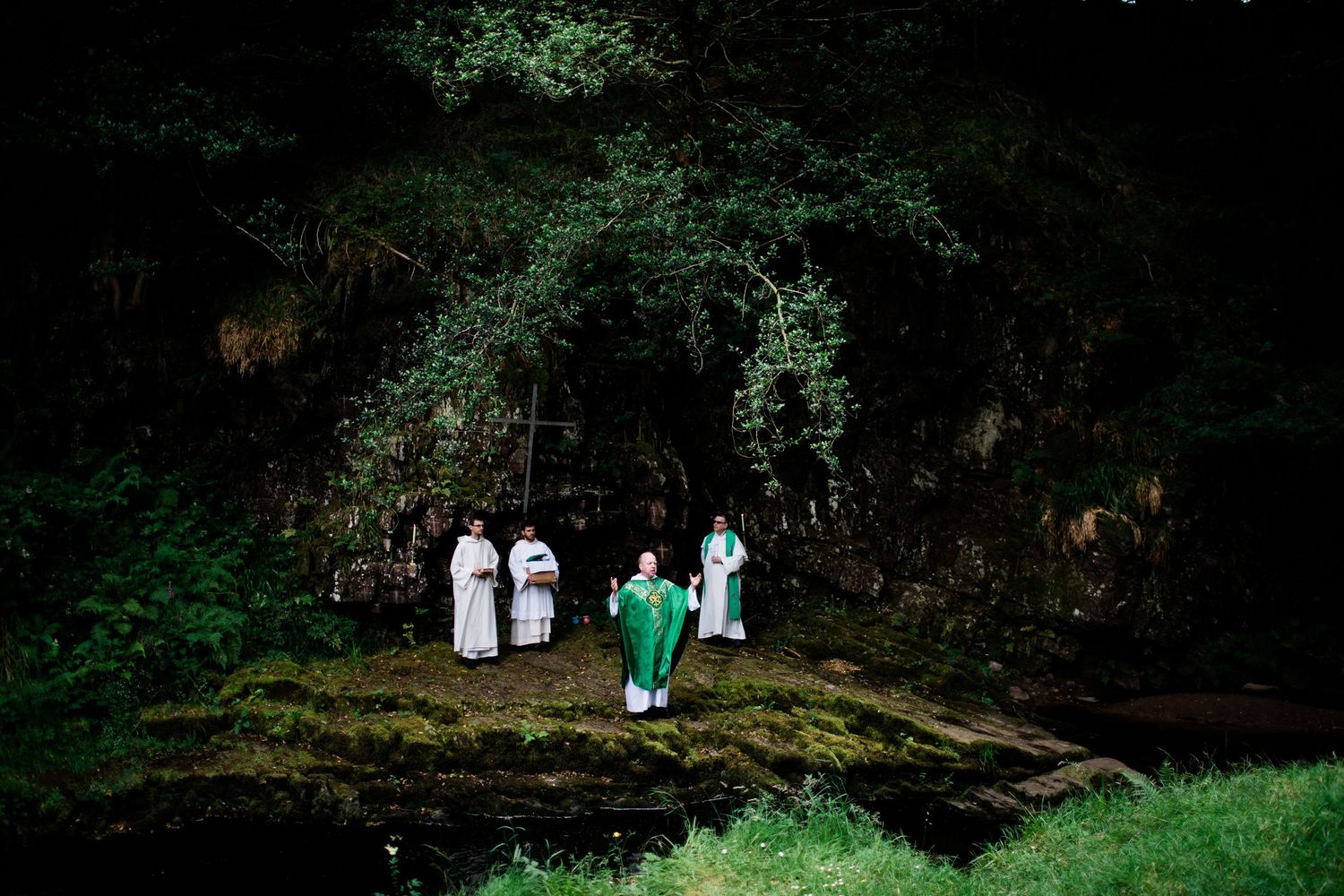 Priests in white and green vestments performing an outdoor religious ceremony near a forest stream.