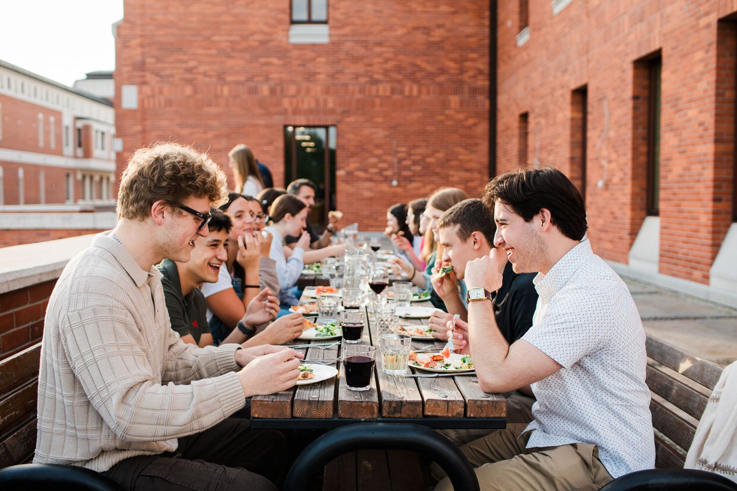 Group of young adults dining together at a long outdoor wooden table against a brick wall.