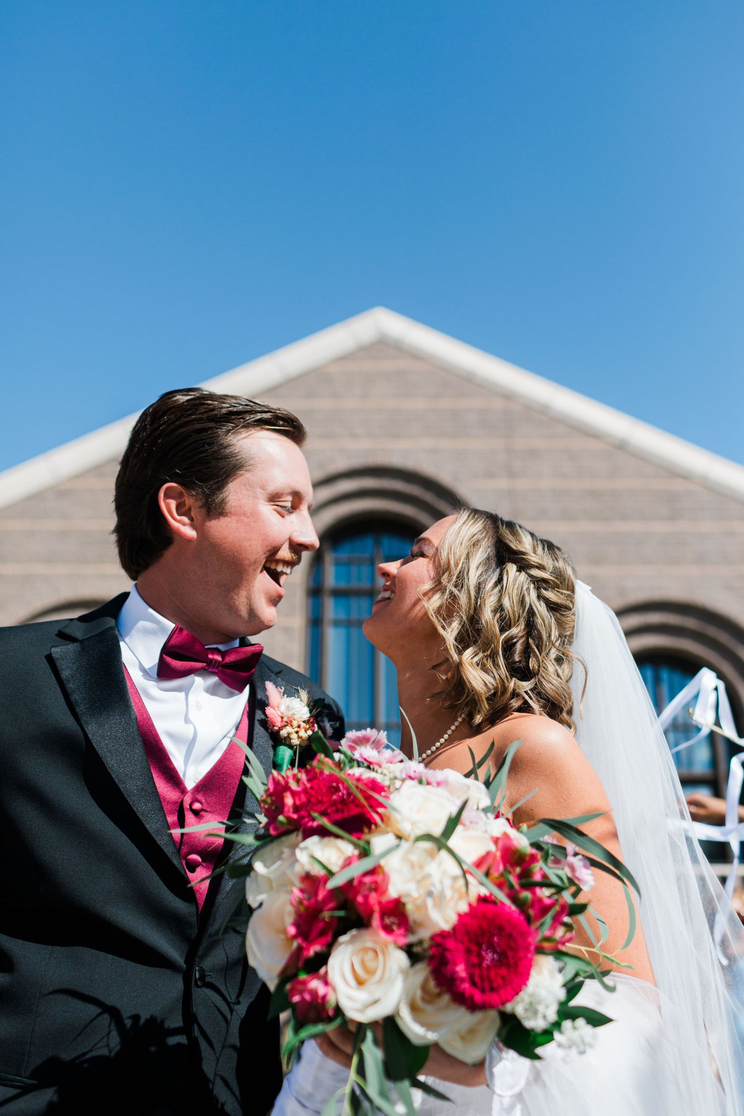 Bride and groom laughing together outside a church on their wedding day, bride holding a bouquet of red and white flowers.
