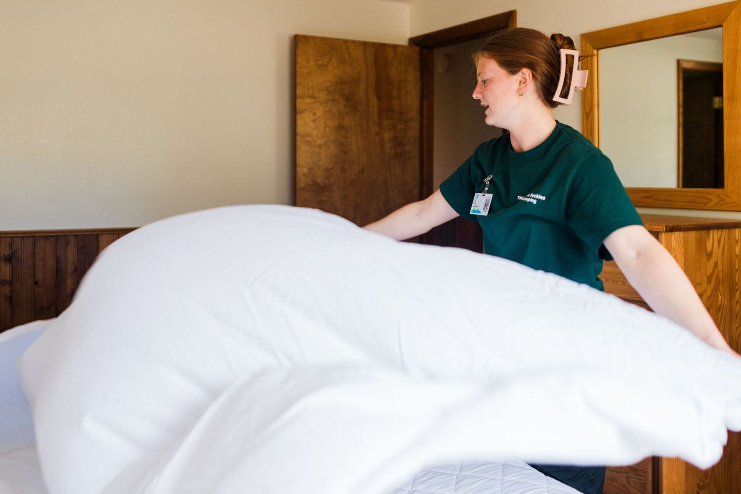 Hotel housekeeper in green uniform making a bed with fresh white linens in a cozy room.