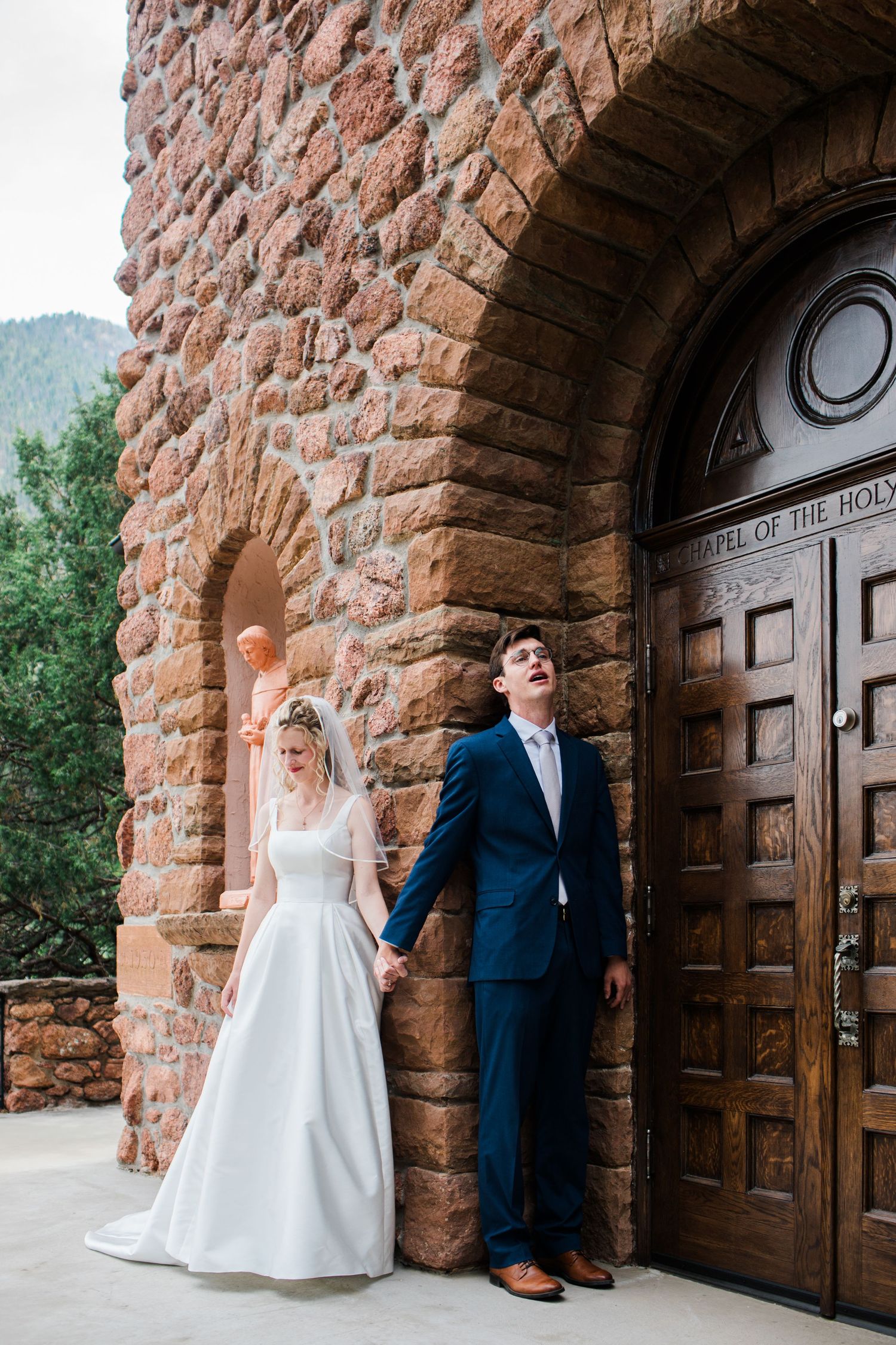Bride and groom hold hands outside Chapel of the Holy Cross stone church, mountains visible in background.