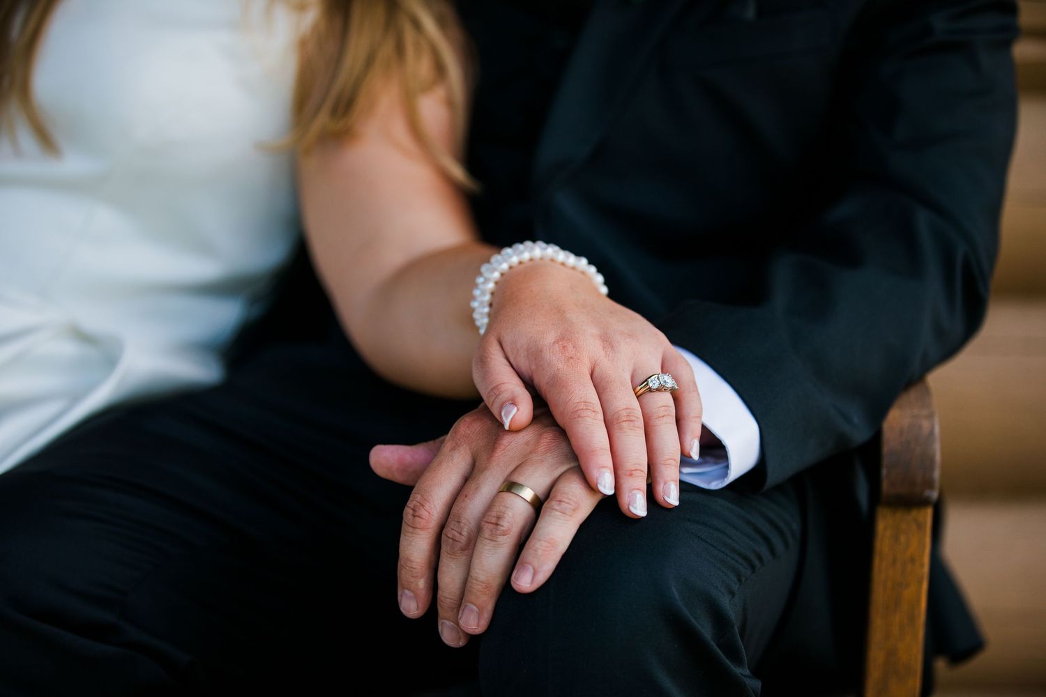 Close-up of newlyweds hands showing wedding rings, bride wearing pearl bracelet, groom in black suit.