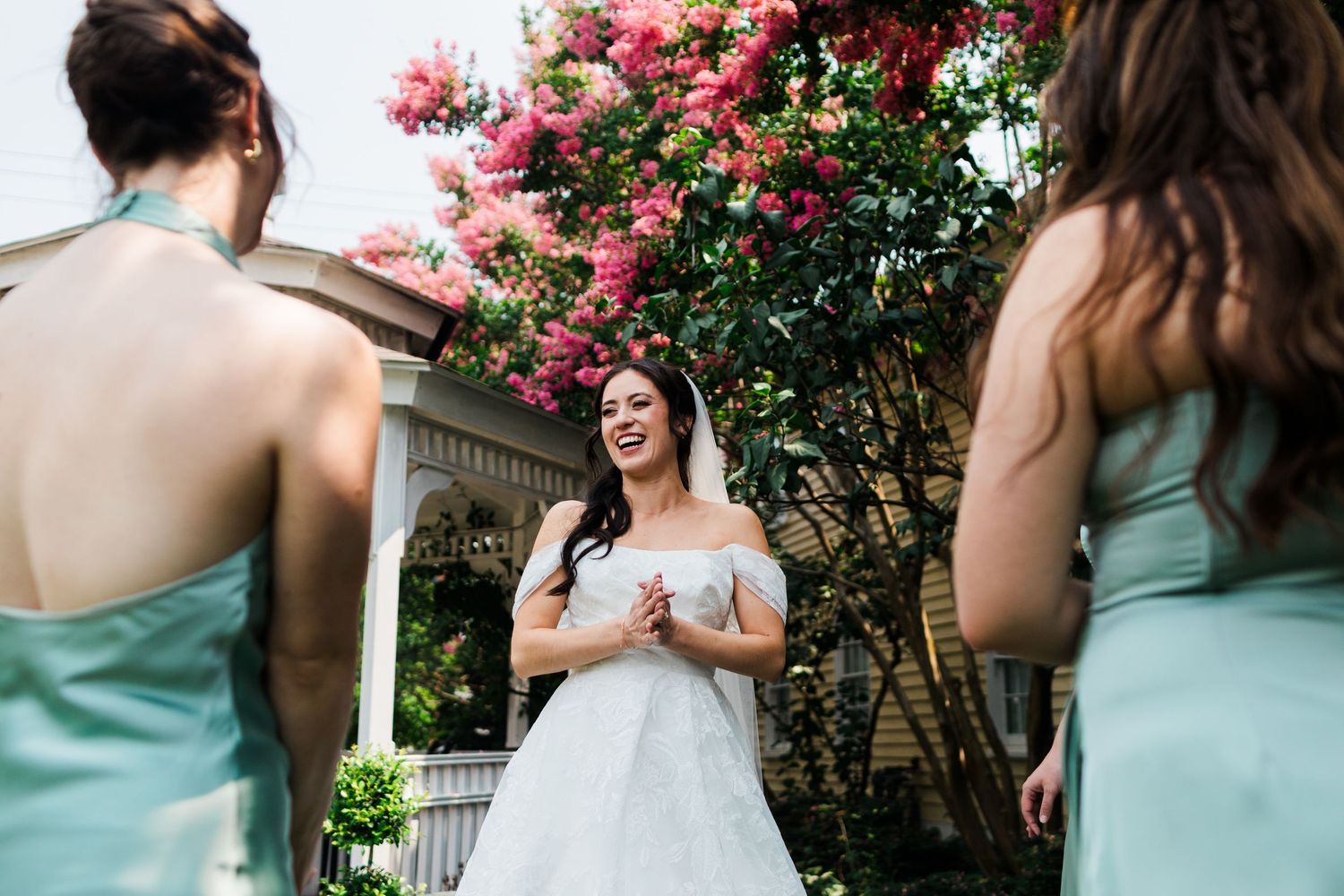 Smiling bride in white gown with bridesmaids in mint dresses near blooming pink flowers.