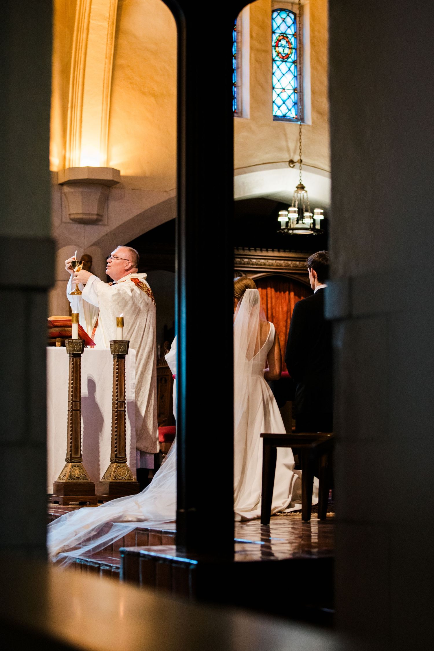 Priest raises chalice during elegant church wedding ceremony as bride in white gown and groom stand at altar.