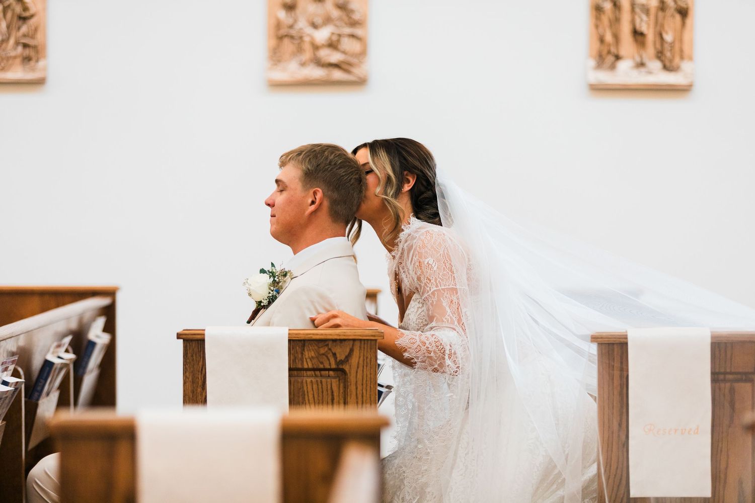 Bride in lace gown and veil sits with groom in white suit during wedding ceremony in a church.