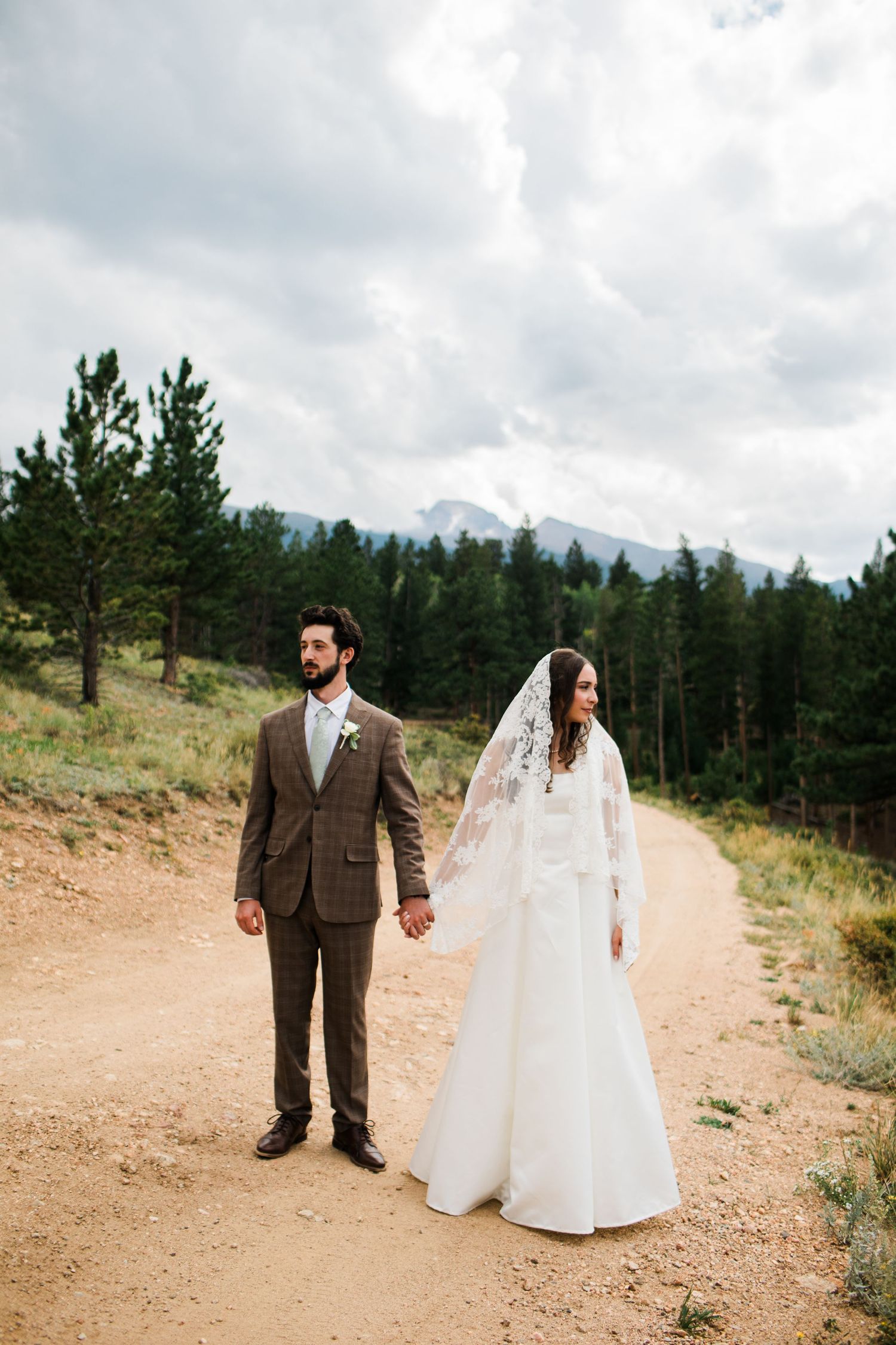 Bride and groom hold hands on a dirt path surrounded by pine trees and mountains on their wedding day.