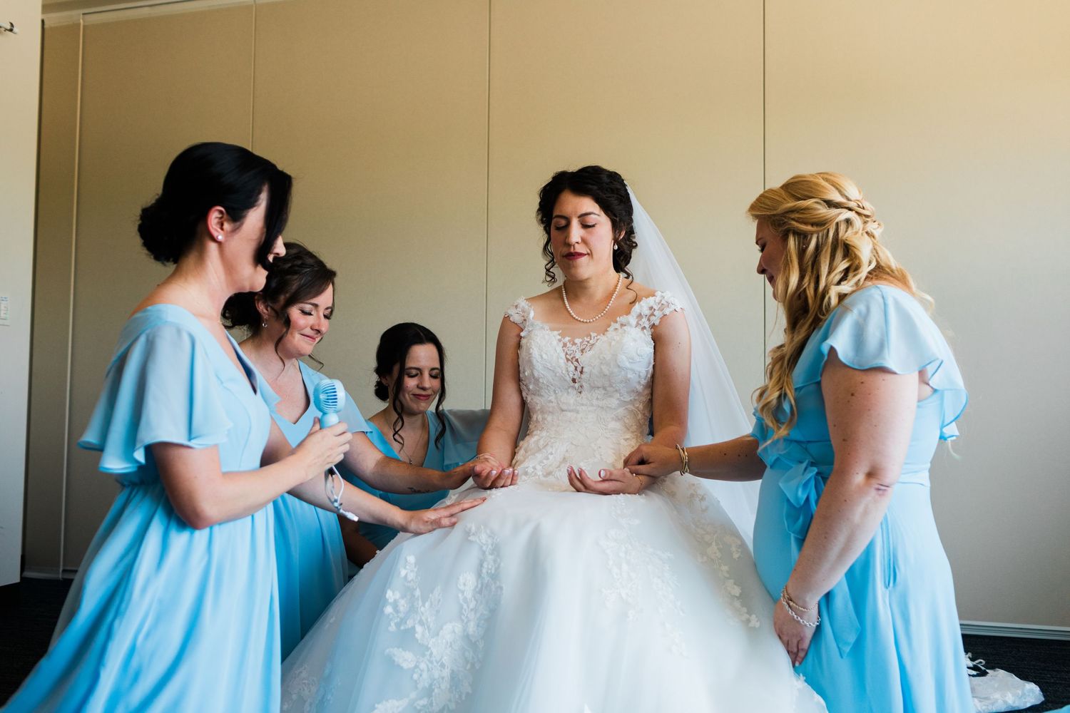 Bride in white lace gown surrounded by bridesmaids in light blue dresses during wedding preparation.