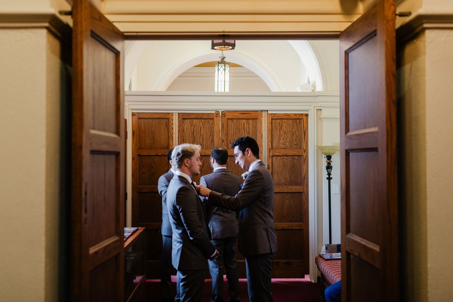 Groomsmen helping each other with ties in a elegant church hallway with wooden doors before a wedding ceremony.