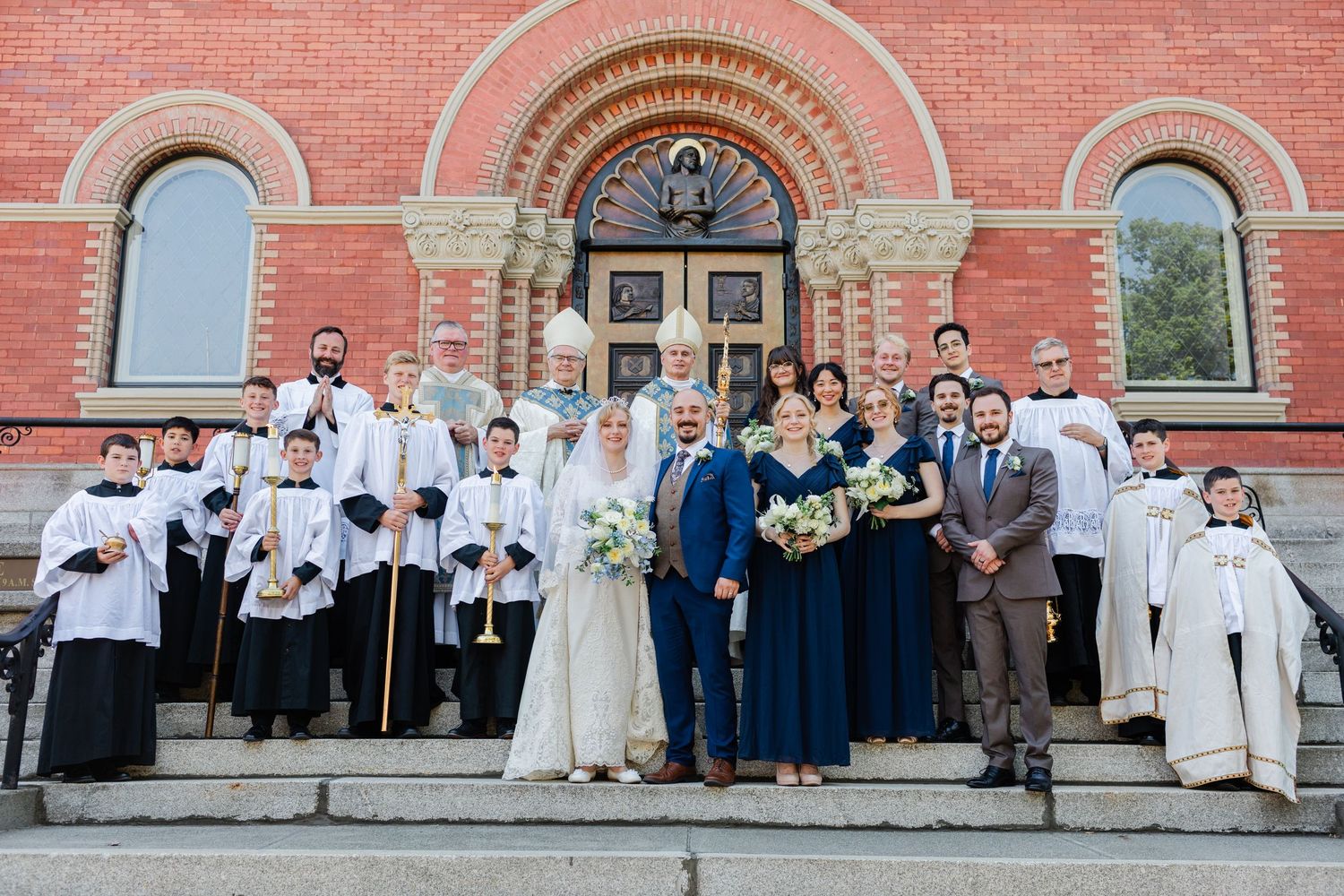 Wedding party poses on church steps with clergy, bride in white, groom in blue suit, and bridesmaids in navy dresses.