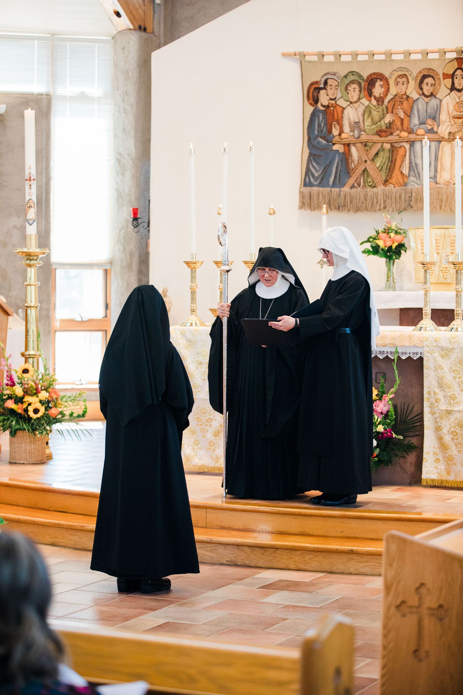 Three nuns in black habits participate in a religious ceremony at a church altar with candles and floral arrangements.