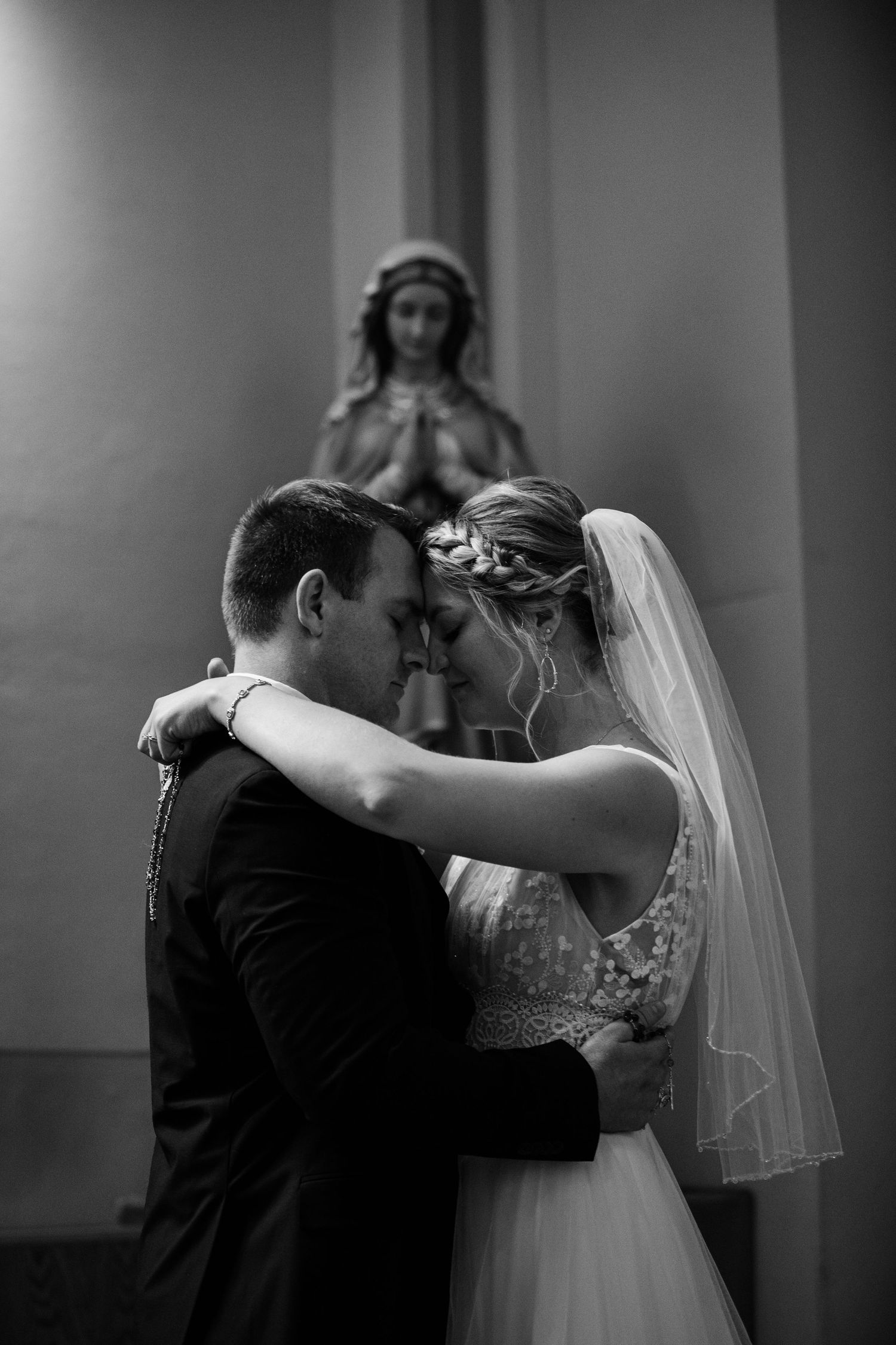 Black and white photo of a bride and groom embracing in a church with a Virgin Mary statue in the background.